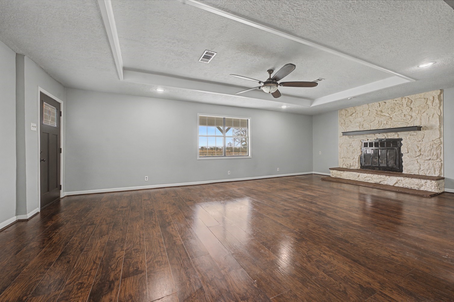 14077 Hamshire Road Hamshire, TX 77622 - Photo 13 of 39 This spacious living room features rich beautiful flooring, a stone fireplace, and a ceiling fan. The room is well-lit with natural light from a large window, offering a cozy yet elegant atmosphere.
