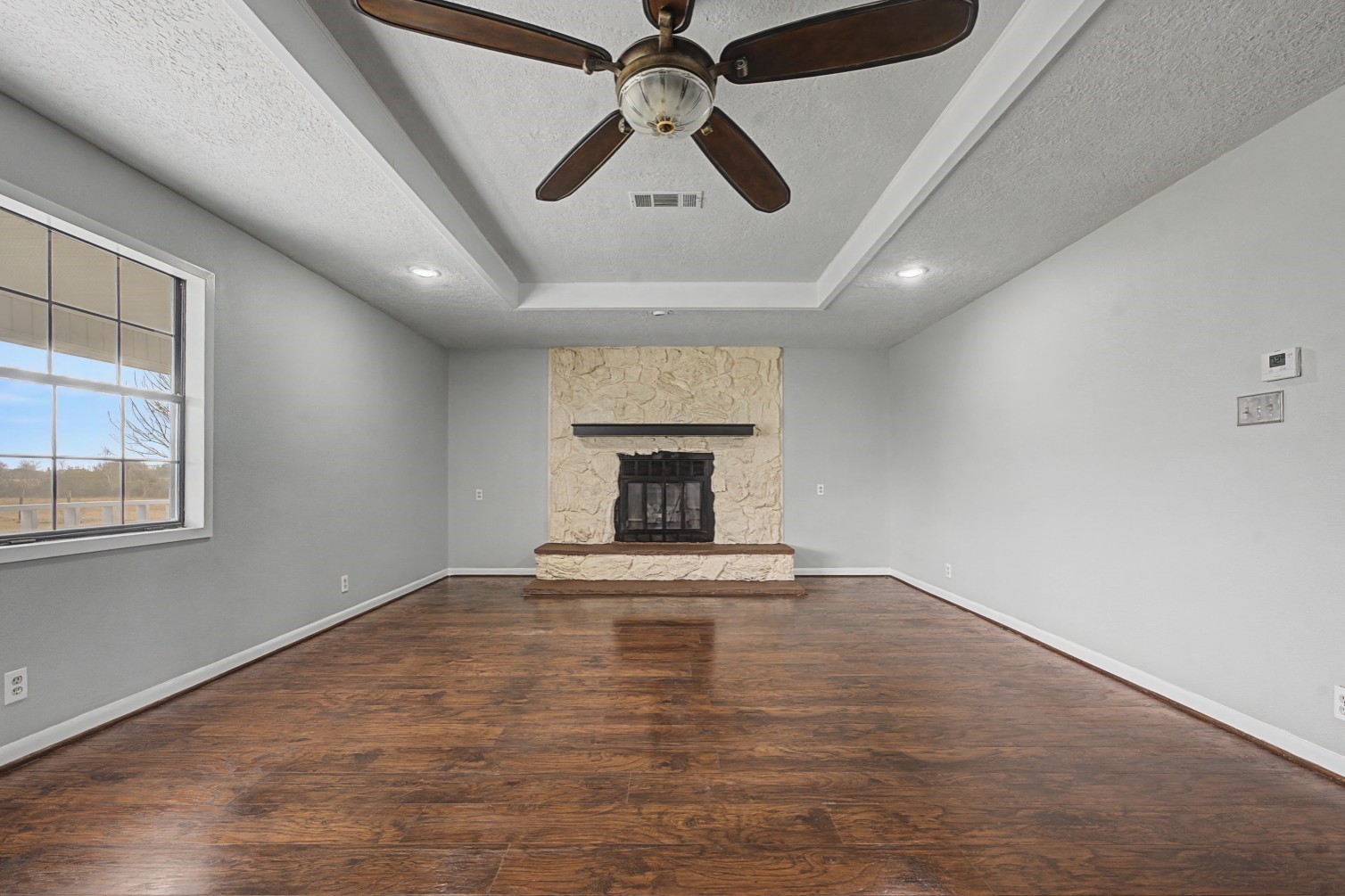 14077 Hamshire Road Hamshire, TX 77622 - Photo 14 of 39 A spacious living room with rich wood like flooring, featuring a stone fireplace as a focal point. The room has a large window for natural light, recessed lighting, and a ceiling fan, creating a cozy and inviting atmosphere.