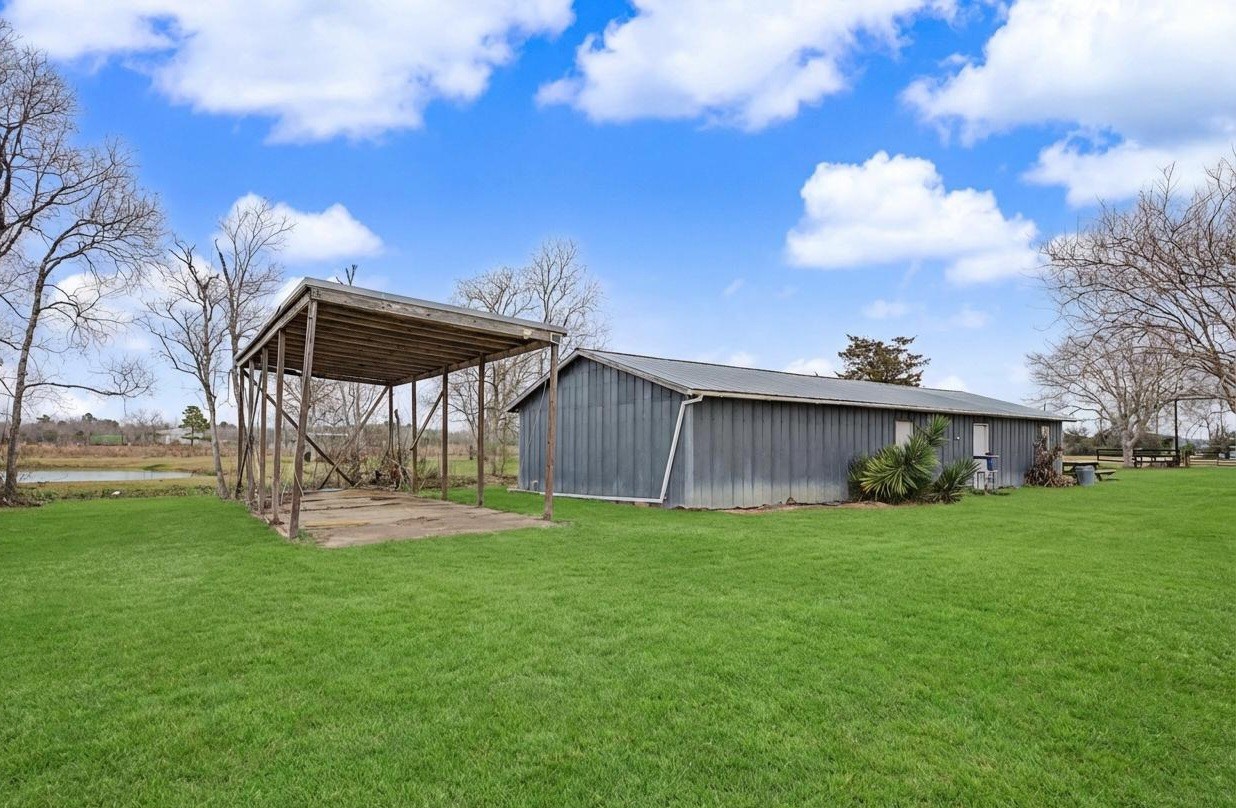 14077 Hamshire Road Hamshire, TX 77622 - Photo 34 of 39 This photo shows a spacious backyard with a 24’x60’ metal storage building and a covered structure. The area is open and grassy, with a few trees and a clear blue sky, offering a rural feel and potential for various outdoor activities.