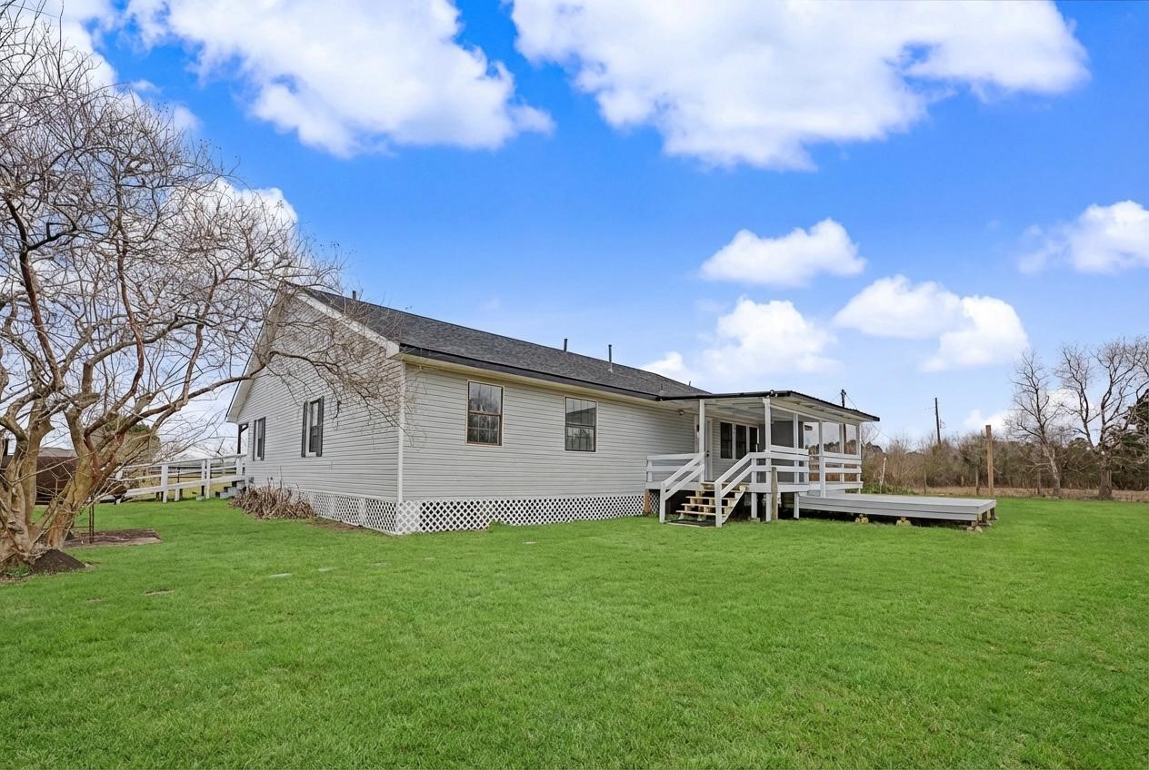 14077 Hamshire Road Hamshire, TX 77622 - Photo 38 of 39 This photo features a single-story house with white siding, a shingled roof, and a spacious wooden deck. The property is surrounded by a large, well-maintained lawn with a few mature trees, all set against a backdrop of clear blue skies.