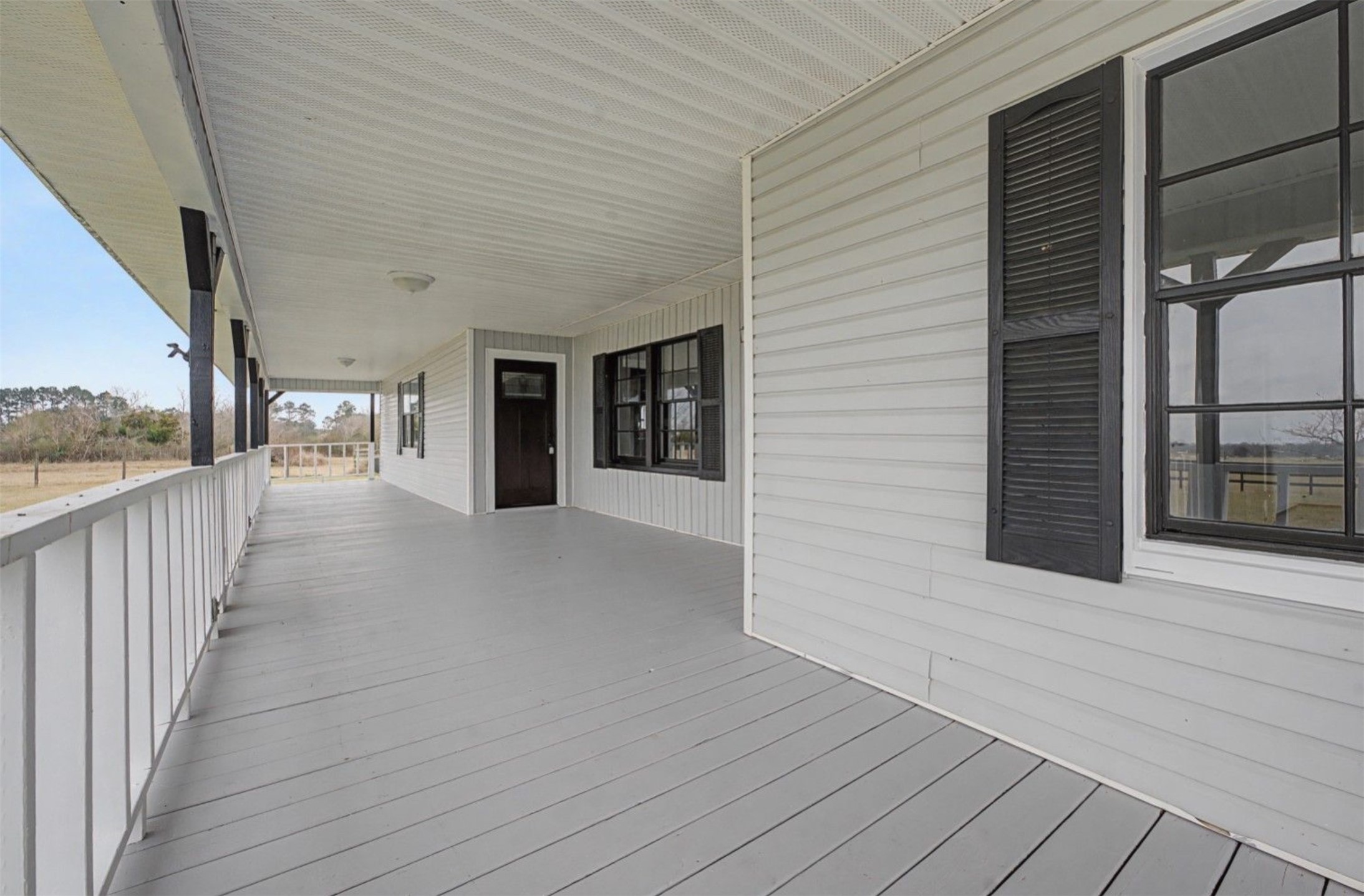 14077 Hamshire Road Hamshire, TX 77622 - Photo 4 of 39 This photo showcases a spacious, covered wrap-around porch with white railings and siding, accented by black shutters. It offers a serene view of open land, ideal for relaxation and enjoying nature.