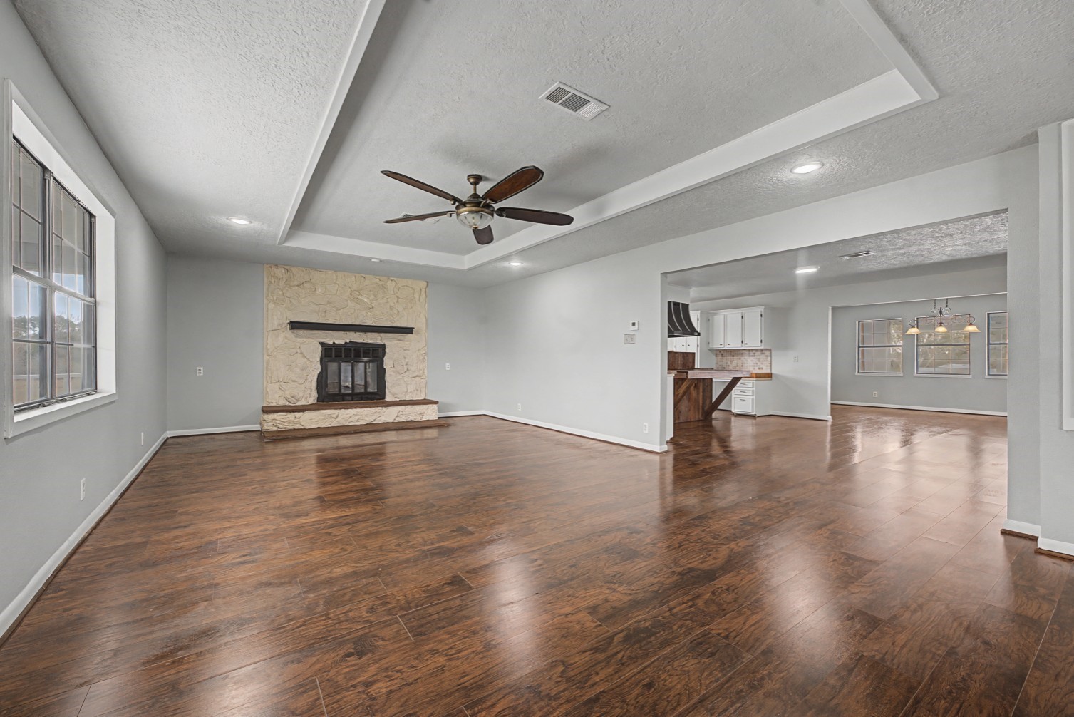 14077 Hamshire Road Hamshire, TX 77622 - Photo 5 of 39 This spacious open-concept living area features warm wood like flooring, a stone fireplace, and a ceiling fan. It seamlessly connects to a modern kitchen with white cabinetry and a dining space, offering ample natural light and a welcoming atmosphere.