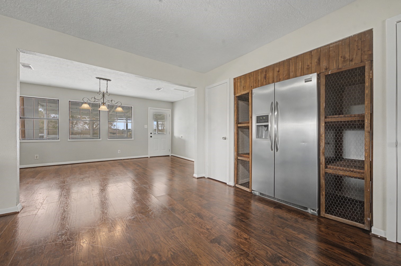 14077 Hamshire Road Hamshire, TX 77622 - Photo 10 of 39 This photo showcases a spacious, open-plan room with polished floors. It features a modern stainless steel fridge set into a rustic wooden cabinet with wire detailing. Large windows provide natural light, and there's a stylish chandelier for added ambiance. The room offers versatile space perfect for dining or living areas.