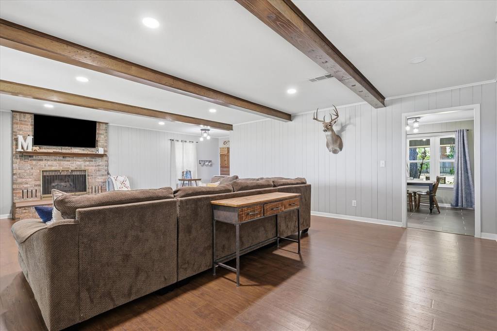 3205 South Garland Street Decatur, TX 76234 - Photo 3 of 30 Spacious living area featuring wood-finish flooring, exposed ceiling beams, a brick fireplace with mantel, recessed lighting, and vertical plank wall paneling