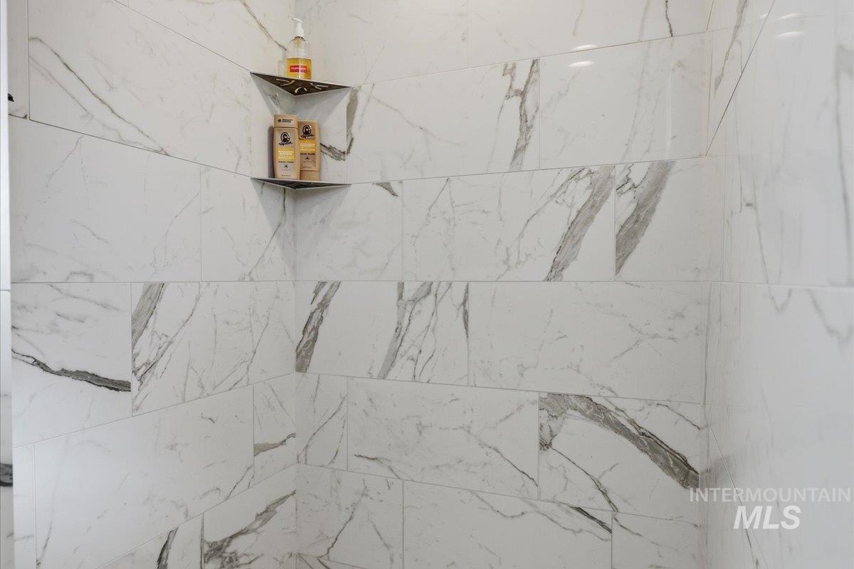 1303 West Brando Street Meridian, ID 83646 - Photo 27 of 49 Shower enclosure featuring large format white tiled walls with gray veining, two built-in corner shelves with dark metallic finishes, and horizontal tile installation