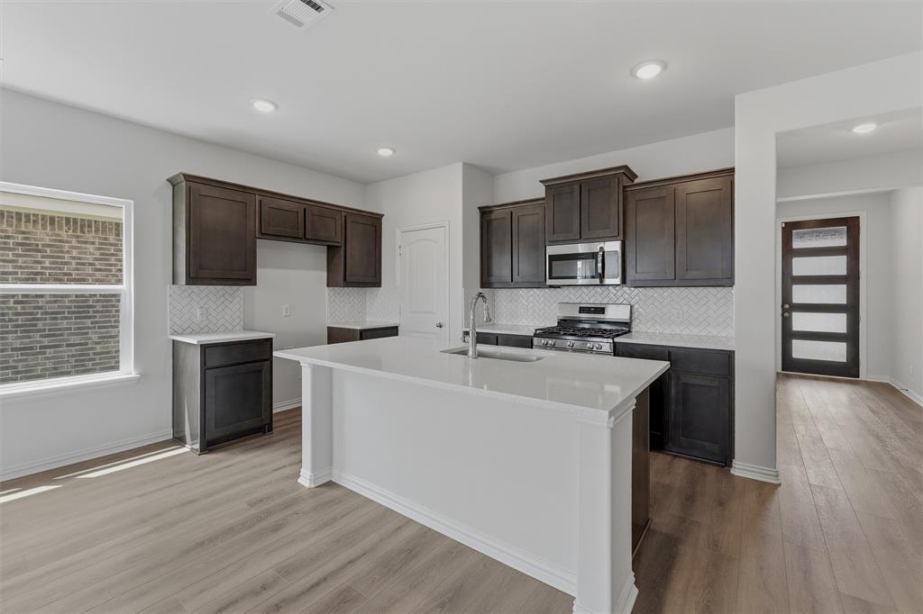 3504 Helena Street Gainesville, TX 76240 - Photo 8 of 19 Contemporary kitchen featuring a large central island with a sink, dark wood-finish cabinetry, stainless steel appliances, a white herringbone tile backsplash, and wood-finish flooring