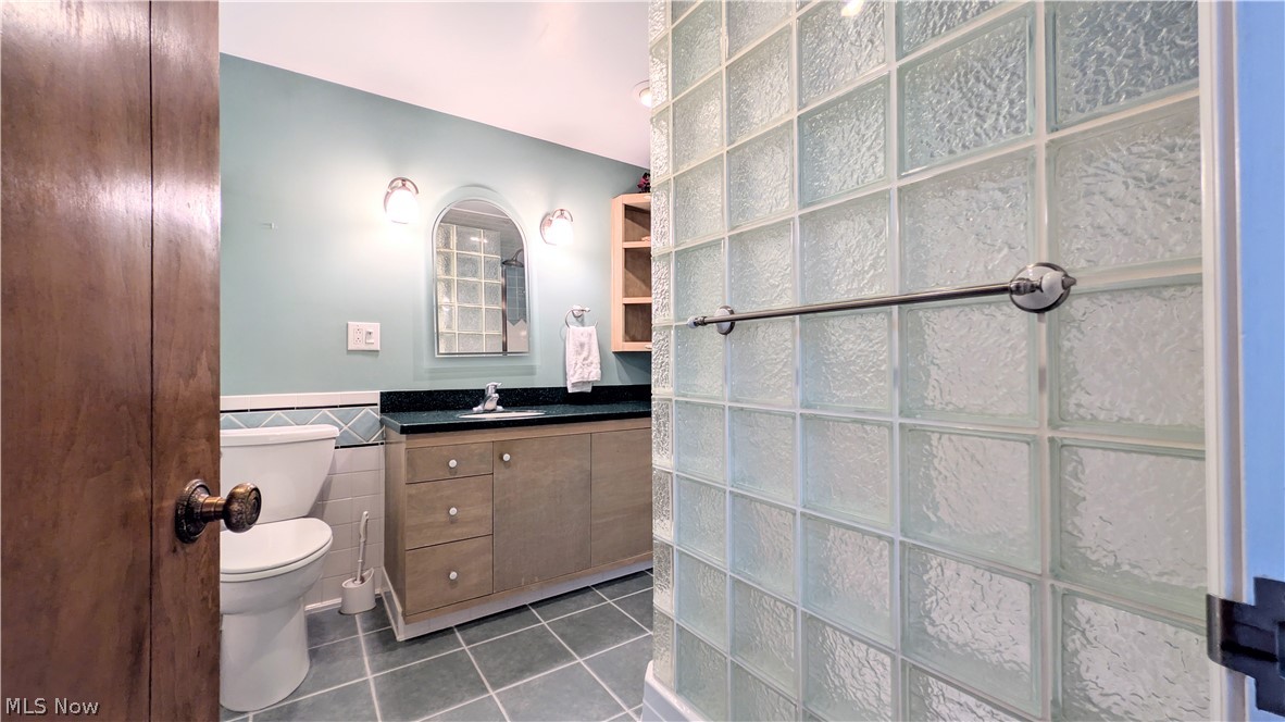 6409 Old Woods Road Ashtabula, OH 44004 - Photo 27 of 43 Bathroom featuring a glass block shower enclosure, wood-finish vanity with a dark countertop, white toilet, and light blue wall tiling