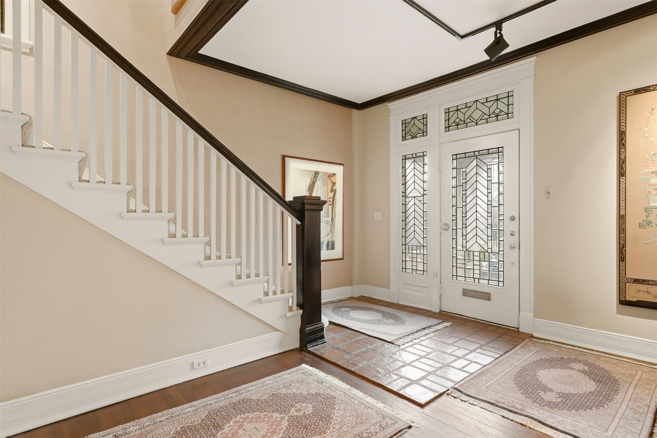 110 Stratford Street Houston, TX 77006 - Photo 9 of 40 This elegant entryway features a staircase with white balusters and a dark wood handrail. The front door showcases decorative glass panels, and the floor is a mix of wood and tiled sections, accented by area rugs. The space is warm and inviting with neutral walls and tasteful artwork.
