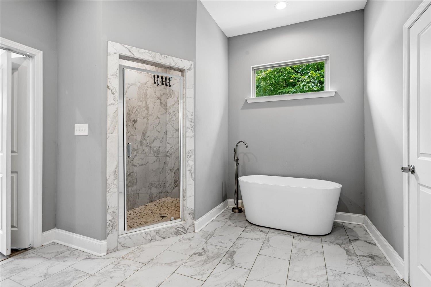 8867 Hampton Court Road East Memphis, TN 38016 - Photo 16 of 23 Spa-inspired primary bathroom featuring a freestanding soaking tub, walk-in shower with marble surround and frameless glass enclosure, marble-finish tile flooring, soft gray walls, recessed ceiling lighting, and a transom window for natural light