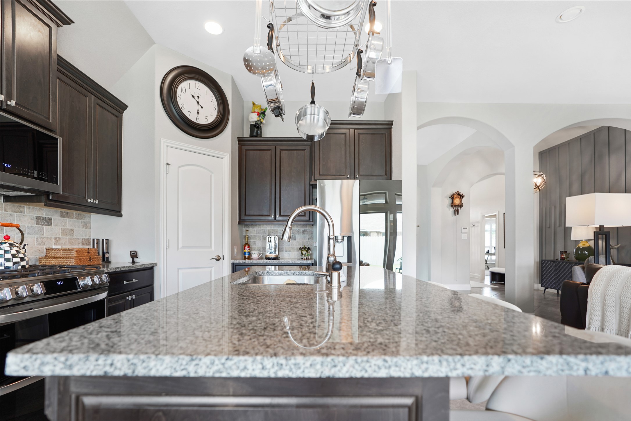 2345 Redwood Ridge Trail Manvel, TX 77578 - Photo 13 of 39 This photo showcases a modern kitchen with elegant dark cabinetry, a granite countertop island, and stainless steel appliances. The open layout flows into a bright living area with stylish décor and arched doorways, creating a welcoming and functional space.