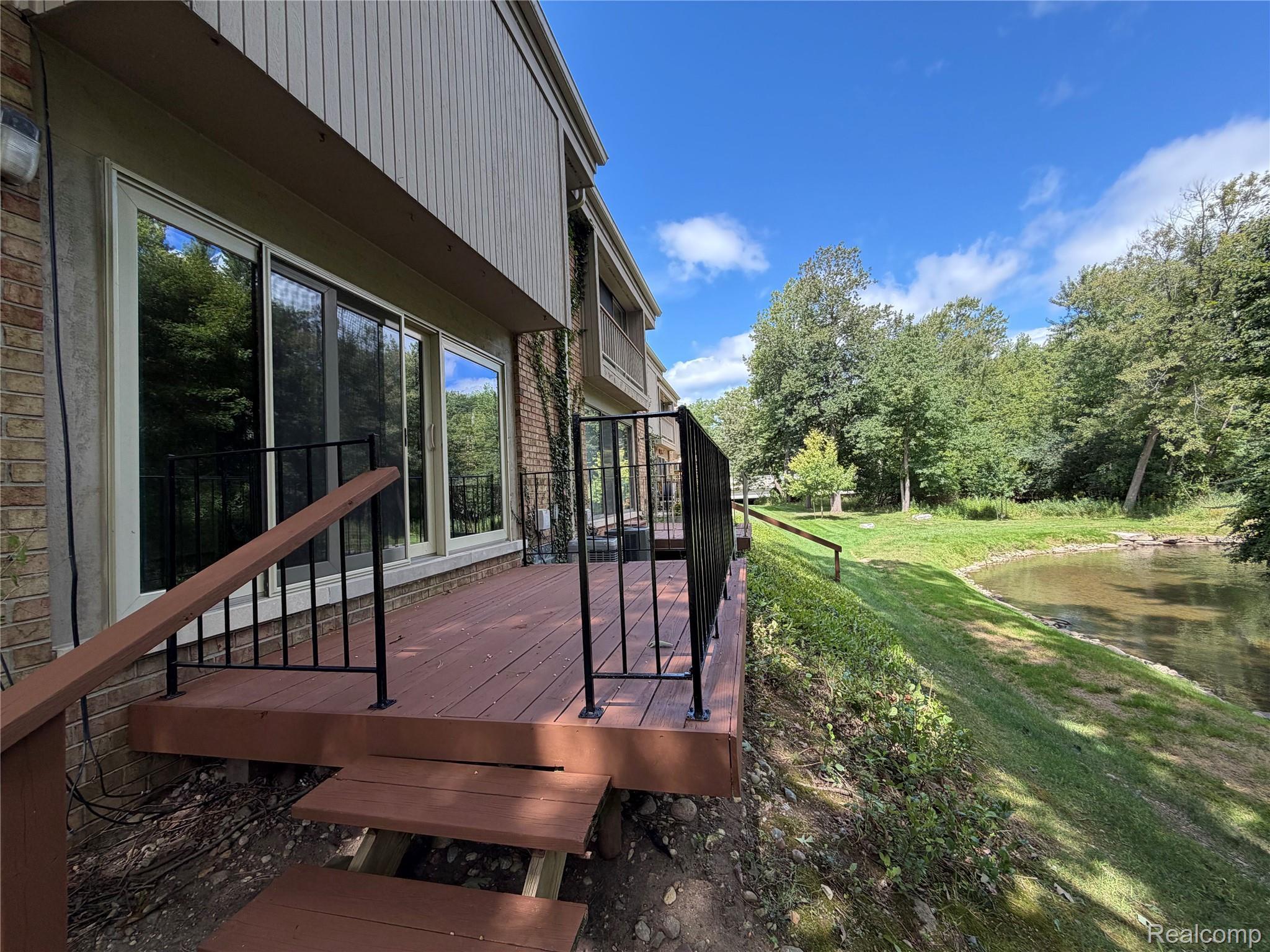 629 Oakbrook West, Unit 18 Rochester, MI 48307 - Photo 5 of 22 Exterior deck with black metal railings, overlooking a grassy embankment, a natural water feature, and mature trees