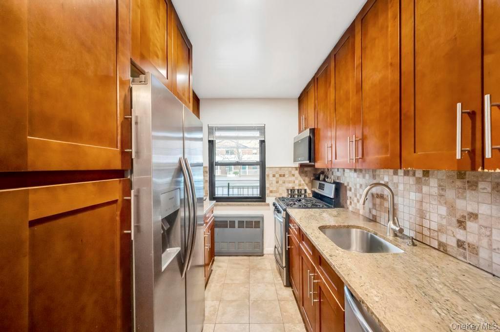 196-72 69th Avenue, Unit 1 Queens, NY 11365 - Photo 10 of 23 Kitchen featuring stainless steel appliances, light stone counters, brown cabinetry, decorative backsplash, and light tile patterned floors
