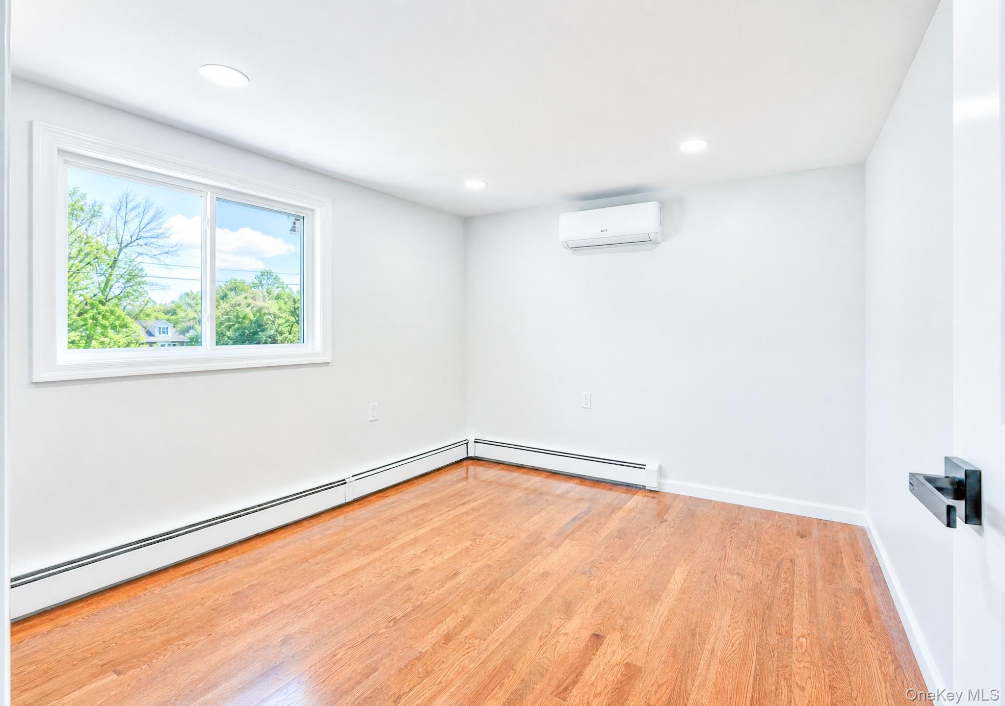 61 Cheshire Road Bethpage, NY 11714 - Photo 13 of 25 Light-filled room featuring wood-finish flooring, a white trim window, baseboard heating, recessed lighting, and a wall-mounted climate control unit