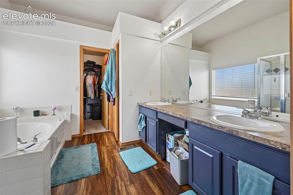 8347 Tibbs Road Peyton, CO 80831 - Photo 17 of 39 Bathroom featuring wood-finish flooring, a built-in tub with white tile surround, a double vanity with dark blue cabinetry, and a walk-in shower with clear glass enclosure
