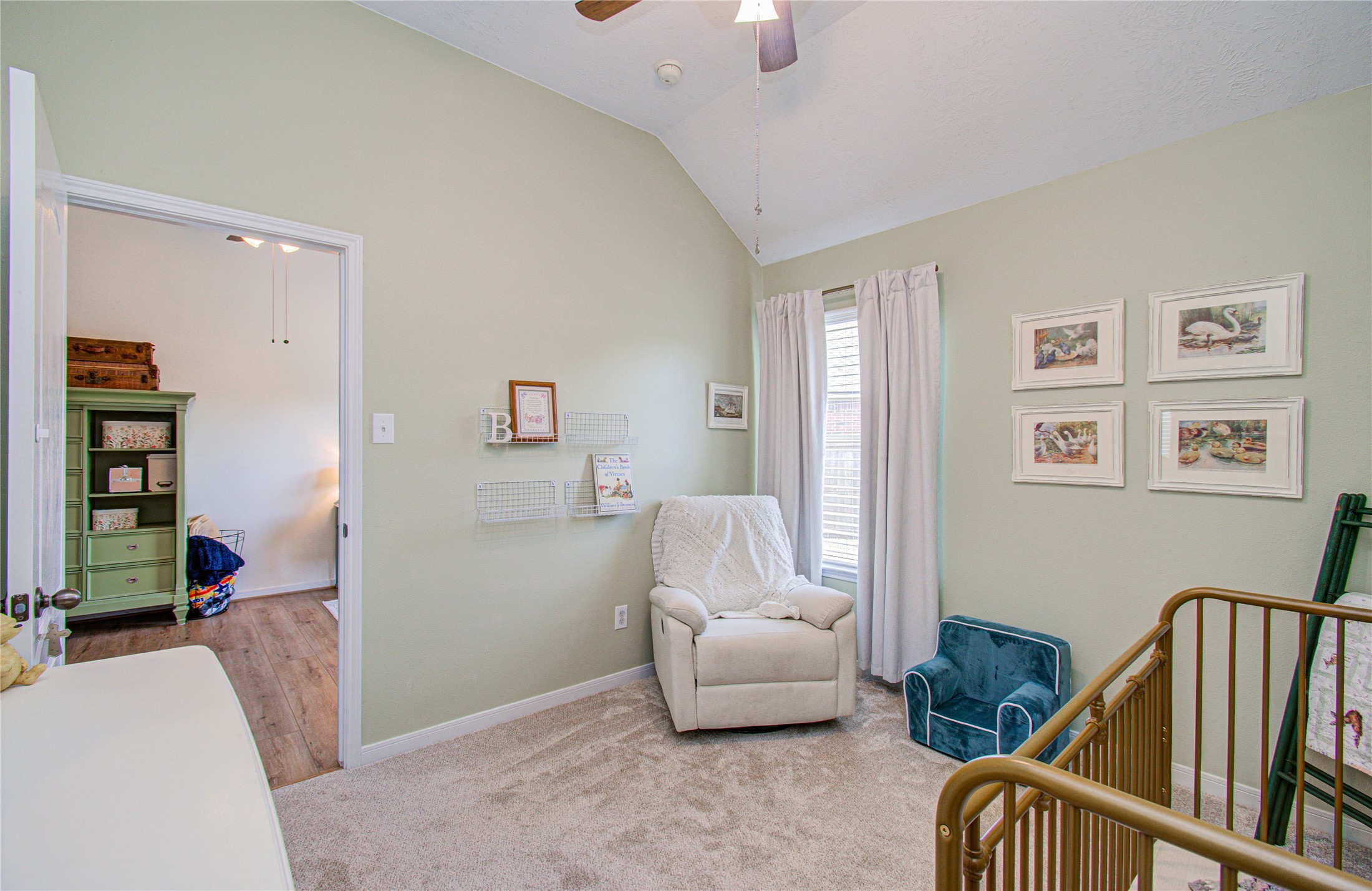 20007 Alcea Court Spring, TX 77379 - Photo 23 of 36 This cozy nursery features soft green walls, carpeted flooring, and a ceiling fan. It includes a comfortable armchair by a bright window with white curtains, wall art, and a crib, creating a welcoming space for a baby.