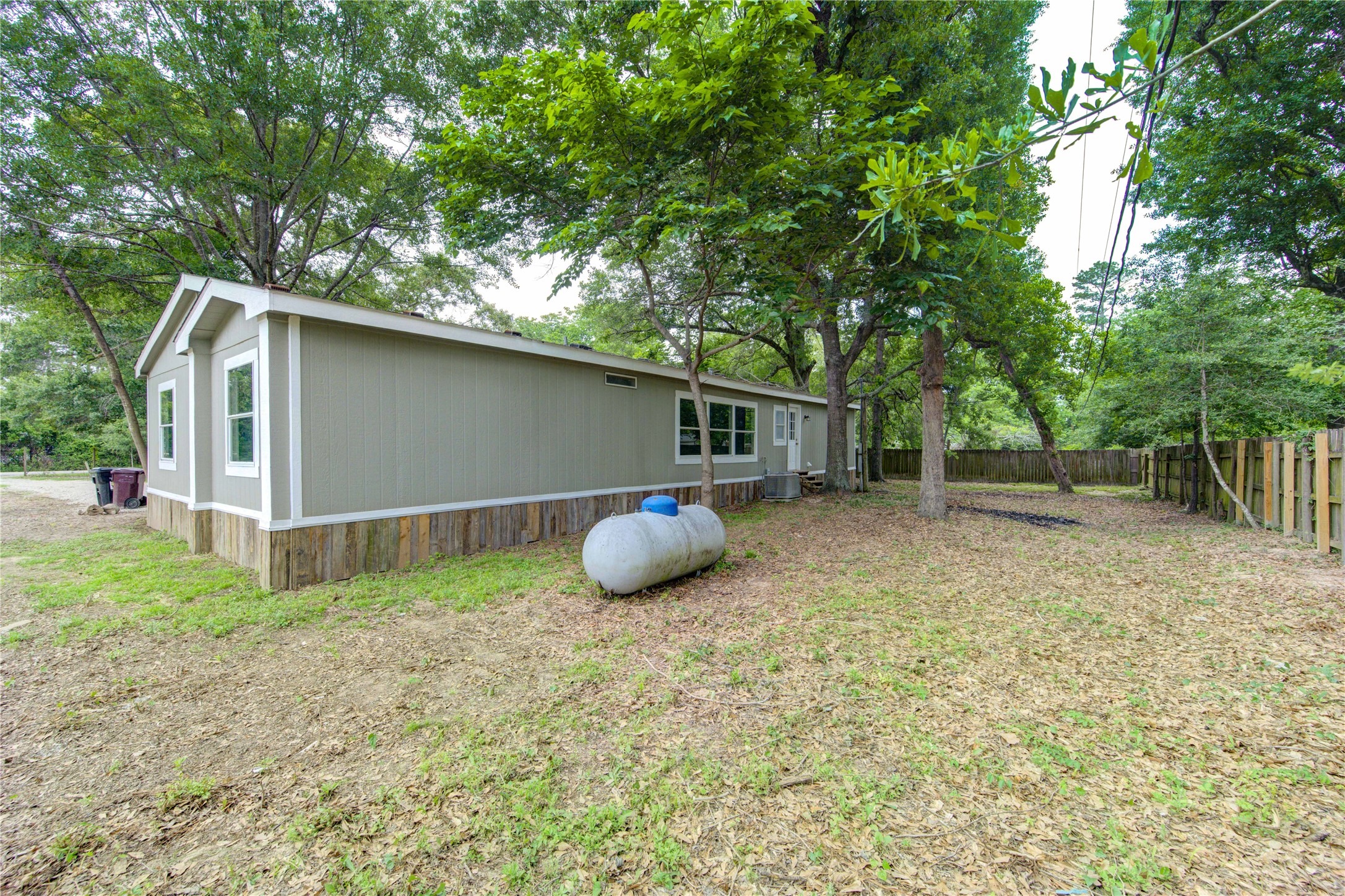 30226 Davis Street Magnolia, TX 77355 - Photo 40 of 50 This photo shows a single-story manufactured home with a light green exterior and white trim. It's set on a spacious, partially wooded lot with a fenced backyard, providing privacy and a natural setting.