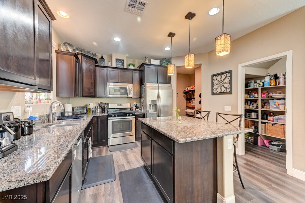 10281 Numaga Road Las Vegas, NV 89178 - Photo 2 of 41 Kitchen with dark brown cabinetry, a kitchen breakfast bar, appliances with stainless steel finishes, light stone counters, and decorative light fixtures