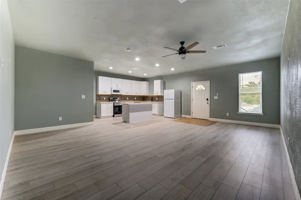 an empty room with wooden floor ceiling fan and kitchen view