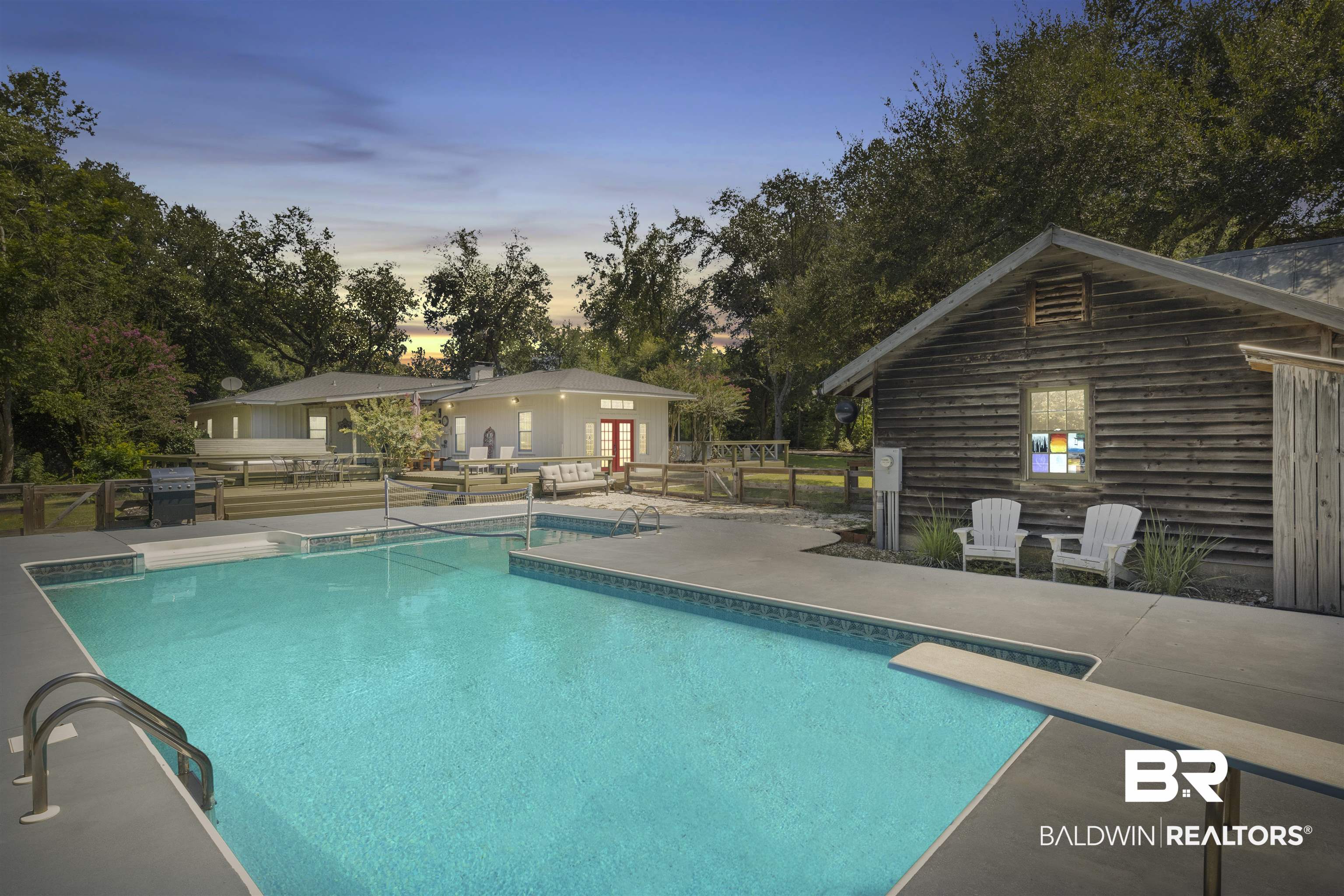 15100 Vasko Road Silverhill, AL 36576 - Photo 2 of 51 Pool at dusk featuring an outdoor structure, a diving board, and a patio next to game/pool cabin. Hot tub, covered porch and outdoor shower are on porch in background.