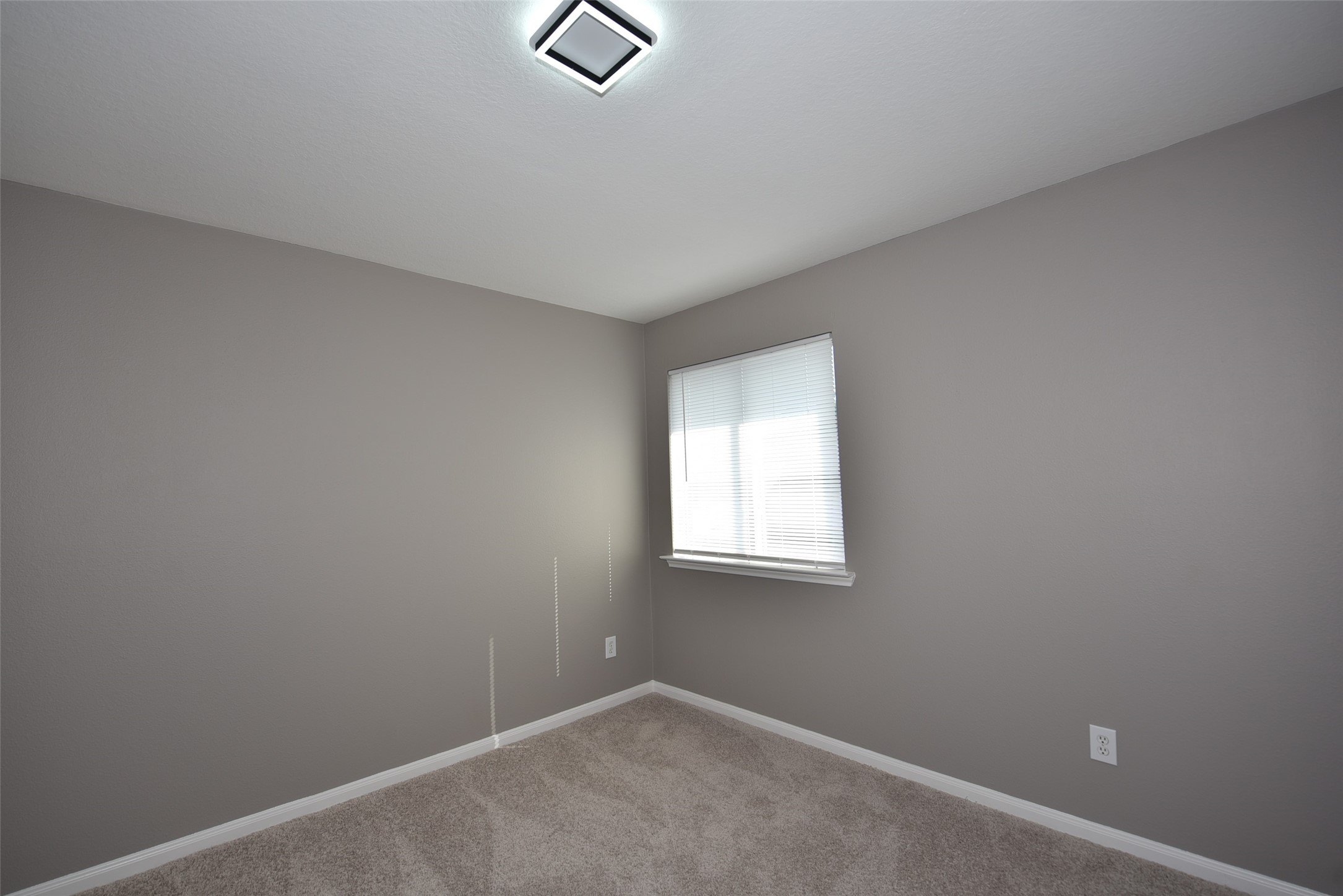 9611 Babbling Spring Court Tomball, TX 77375 - Photo 24 of 31 This photo shows a small, simple room with neutral gray walls and a light carpet. It features a window with blinds that allow natural light, and a modern ceiling light fixture. Ideal for a cozy bedroom or home office.