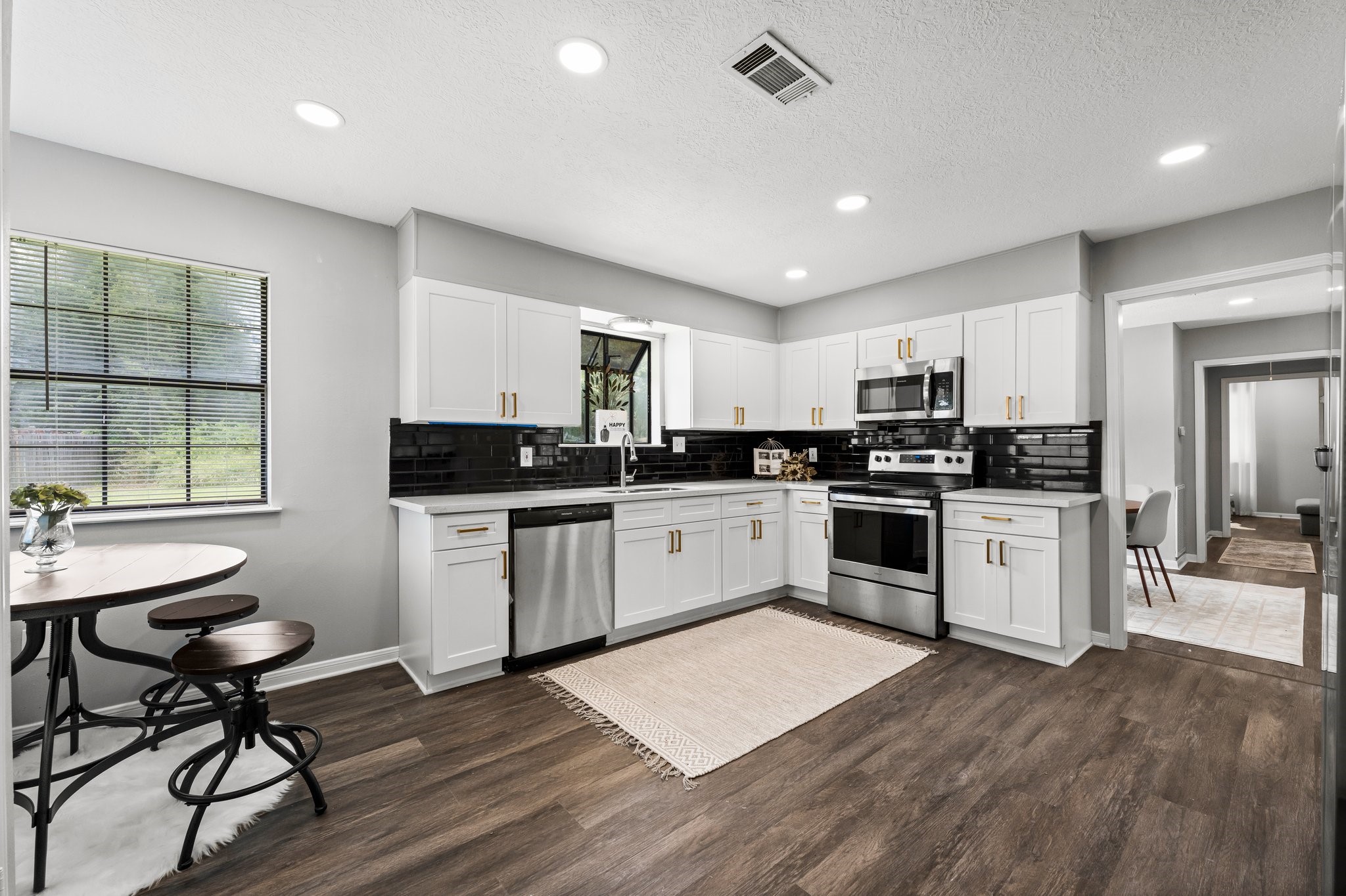 7280 Click Drive Beaumont, TX 77708 - Photo 10 of 39 This modern kitchen features sleek white cabinetry with gold handles, stainless steel appliances, and a stylish black tile backsplash. The room is well-lit with recessed lighting and has a cozy breakfast nook by the window, offering a pleasant view of the outdoors. The dark wood flooring adds warmth and contrast to the space.