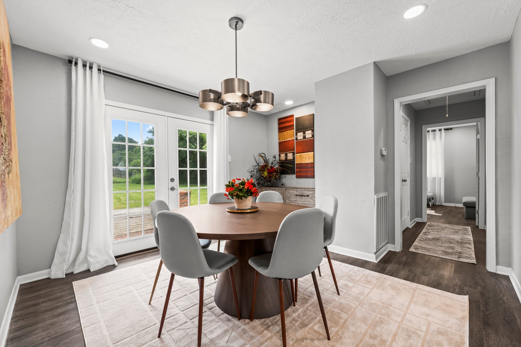 7280 Click Drive Beaumont, TX 77708 - Photo 13 of 39 This photo showcases a bright, modern dining area with a round table and stylish chairs. It features sleek wood flooring, contemporary lighting, and large glass doors that lead to a lush backyard, providing ample natural light. The neutral color palette creates a welcoming atmosphere, perfect for entertaining.