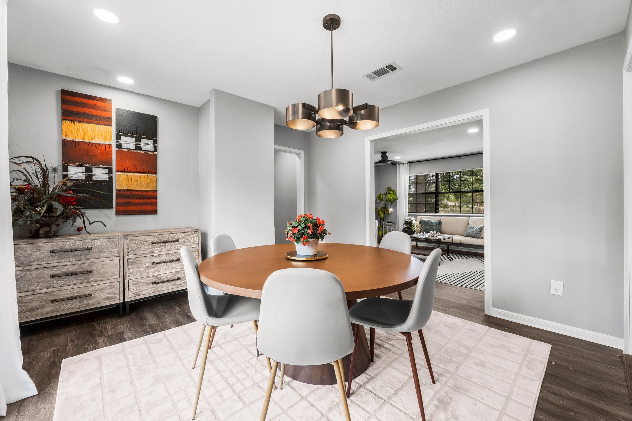 7280 Click Drive Beaumont, TX 77708 - Photo 14 of 39 This photo showcases a modern dining area with a round wooden table and stylish chairs on a light rug. The room features a contemporary light fixture and neutral walls, complemented by decorative artwork and a sideboard. A glimpse into the adjacent living area reveals large windows that bring in natural light.