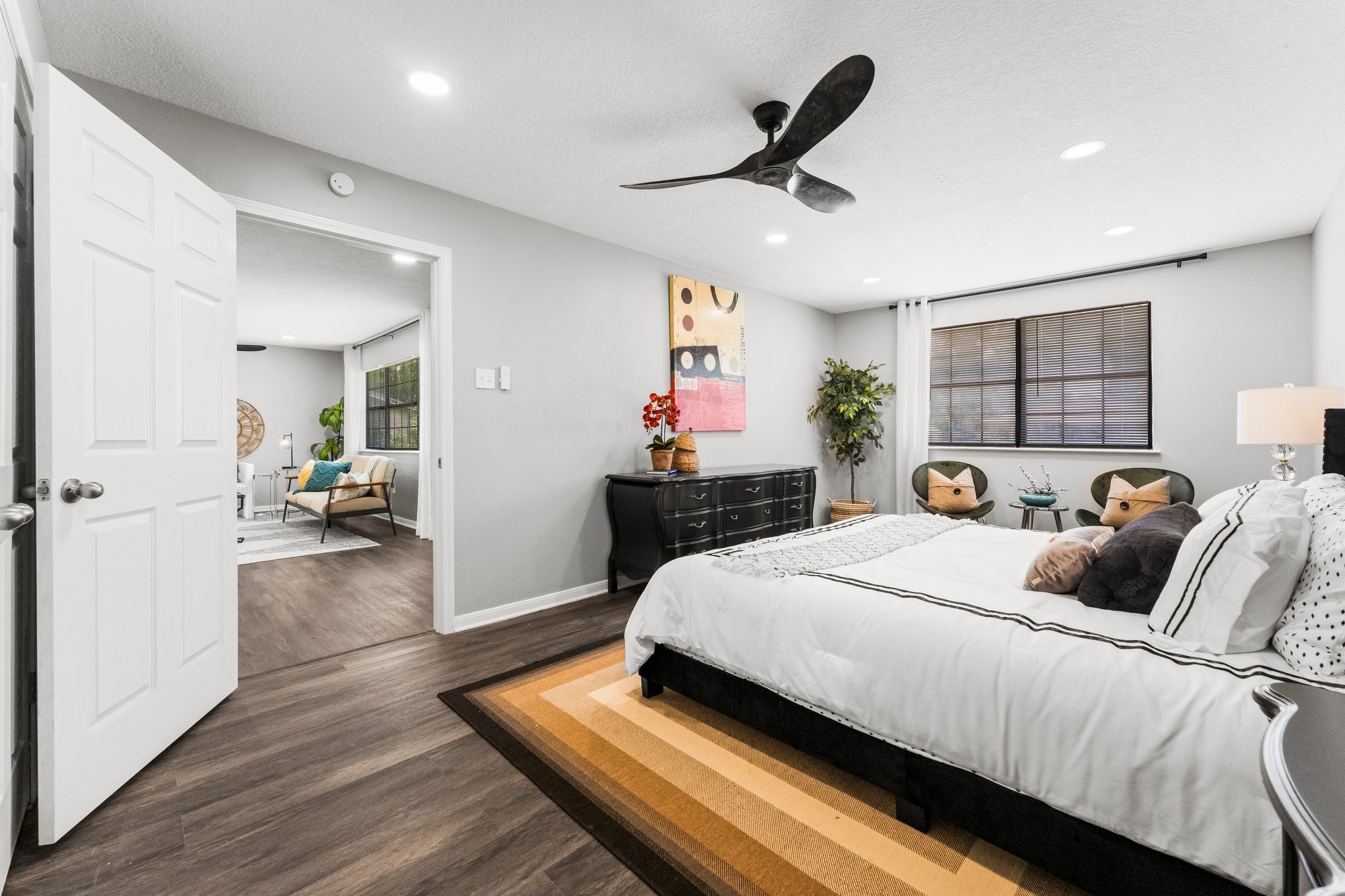 7280 Click Drive Beaumont, TX 77708 - Photo 17 of 39 This photo showcases a modern, spacious bedroom with sleek hardwood floors and a large window for natural light. The room features a stylish ceiling fan, contemporary furniture, and neutral colors for a calm atmosphere. An open door leads to an adjacent living area, enhancing the flow and connectivity of the space.