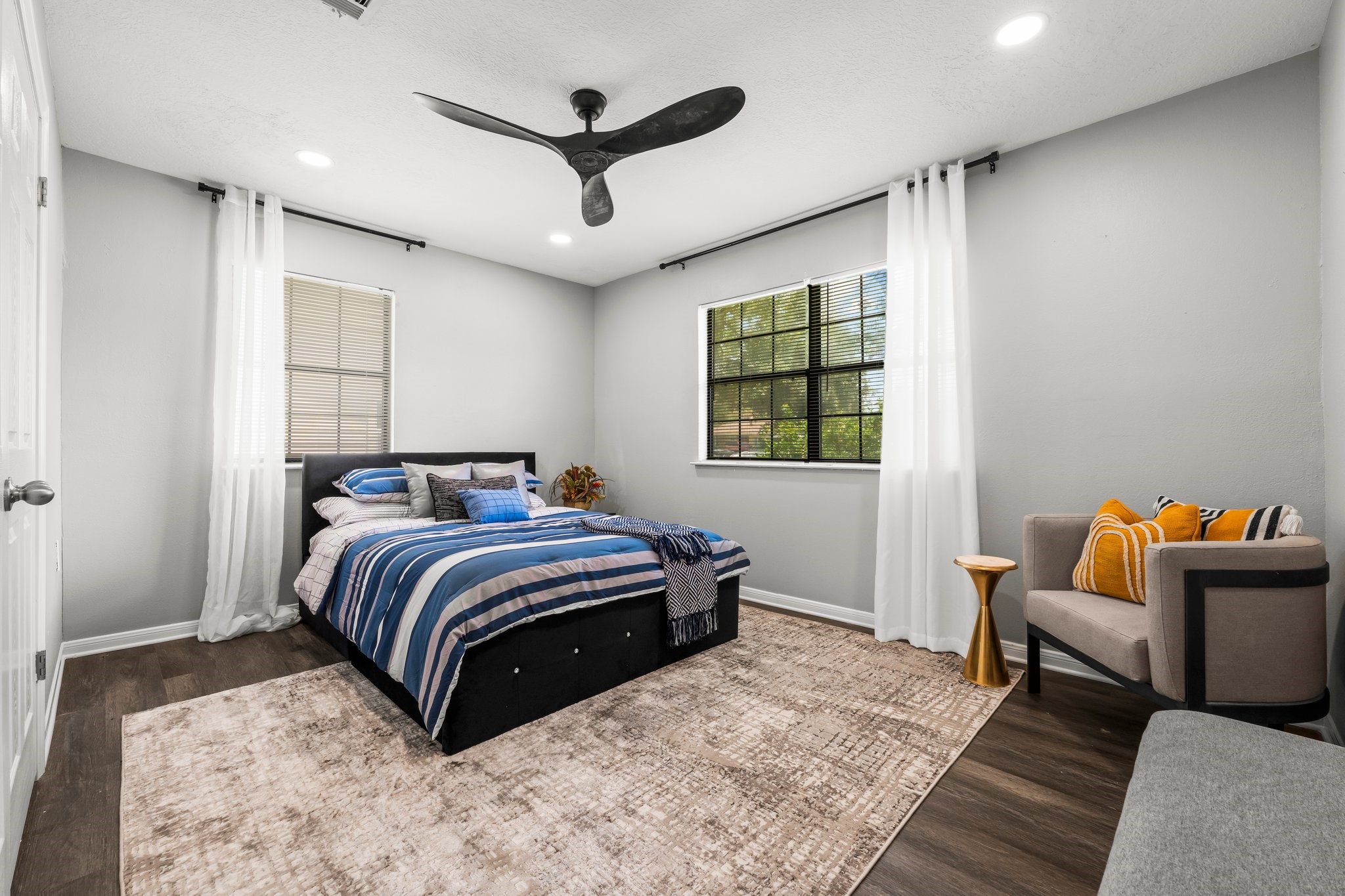 7280 Click Drive Beaumont, TX 77708 - Photo 18 of 39 This bedroom features a modern design with a cozy bed, stylish ceiling fan, and wood flooring. Large windows with sheer curtains provide natural light, complemented by a comfortable chair and decorative accents, creating a welcoming and serene atmosphere.