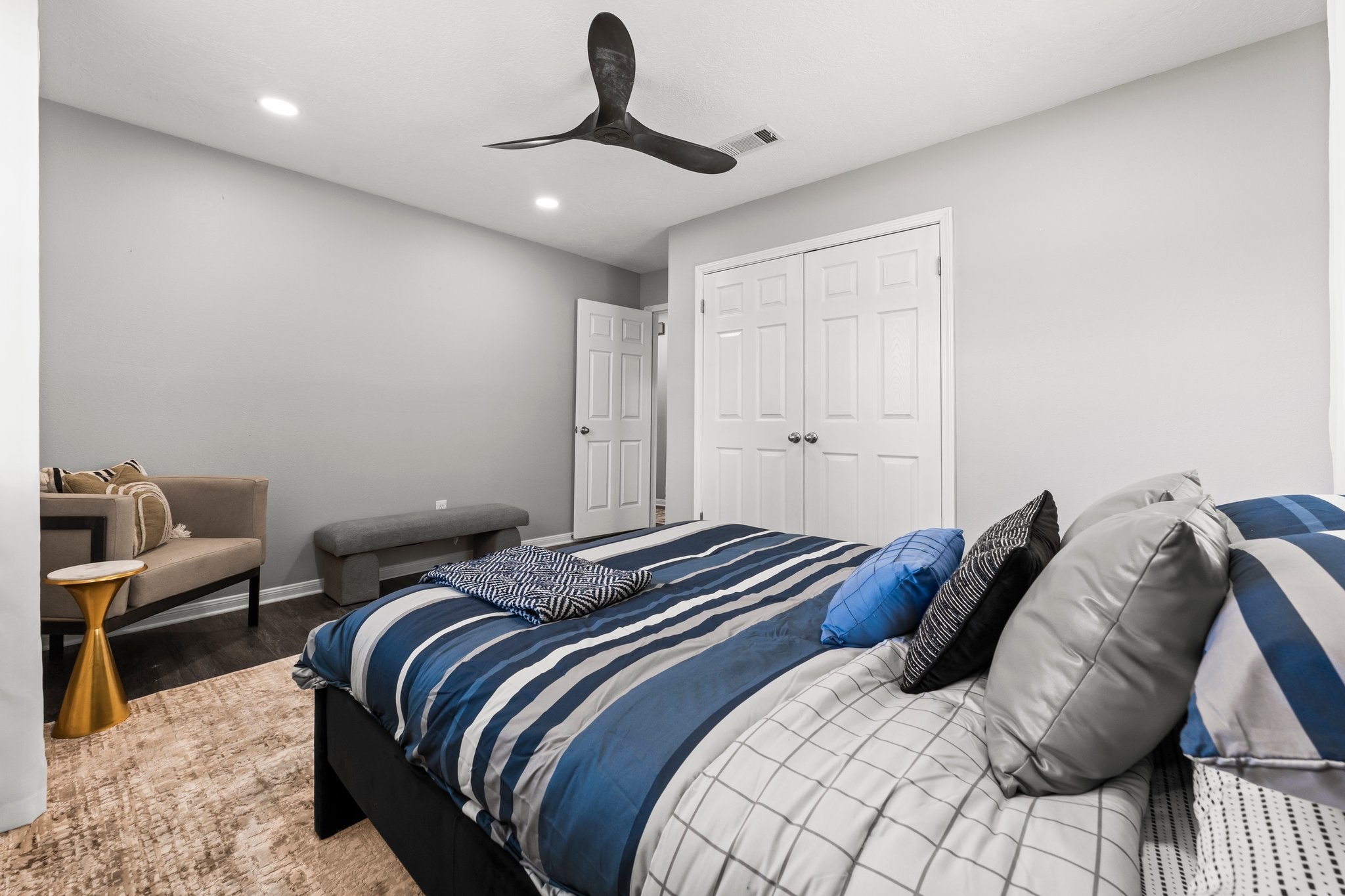 7280 Click Drive Beaumont, TX 77708 - Photo 19 of 39 This bedroom features a modern design with neutral walls, a ceiling fan, and recessed lighting. It includes a comfortable bed with striped bedding, a cozy seating area with a chair and bench, and a spacious closet with double doors. The room has dark flooring and a stylish accent table.