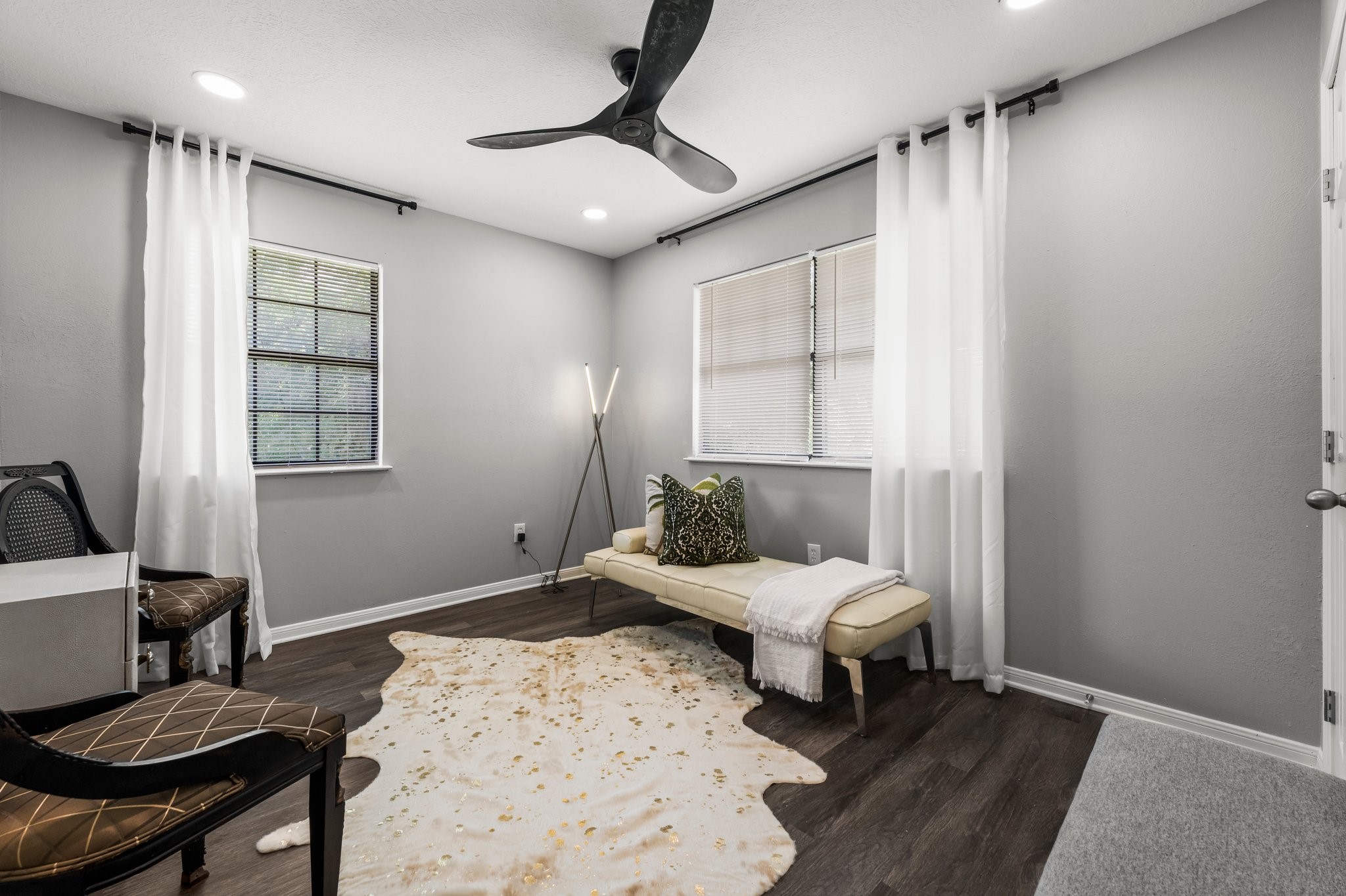 7280 Click Drive Beaumont, TX 77708 - Photo 20 of 39 A cozy, modern room with gray walls and dark wood flooring. It features a ceiling fan, ample natural light from two windows with white curtains, and stylish furnishings, including a bench and two chairs. A decorative rug and floor lamp add a touch of elegance.