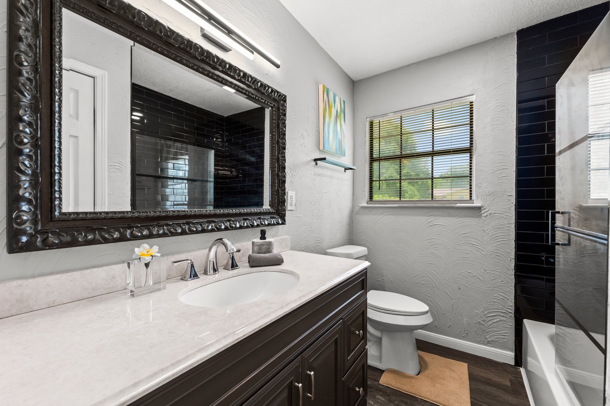 7280 Click Drive Beaumont, TX 77708 - Photo 22 of 39 This bathroom features a modern design with a sleek black vanity and a large decorative mirror. It includes a spacious countertop, elegant fixtures, and a glass-enclosed shower. The window provides natural light, enhancing the room's bright and airy feel.