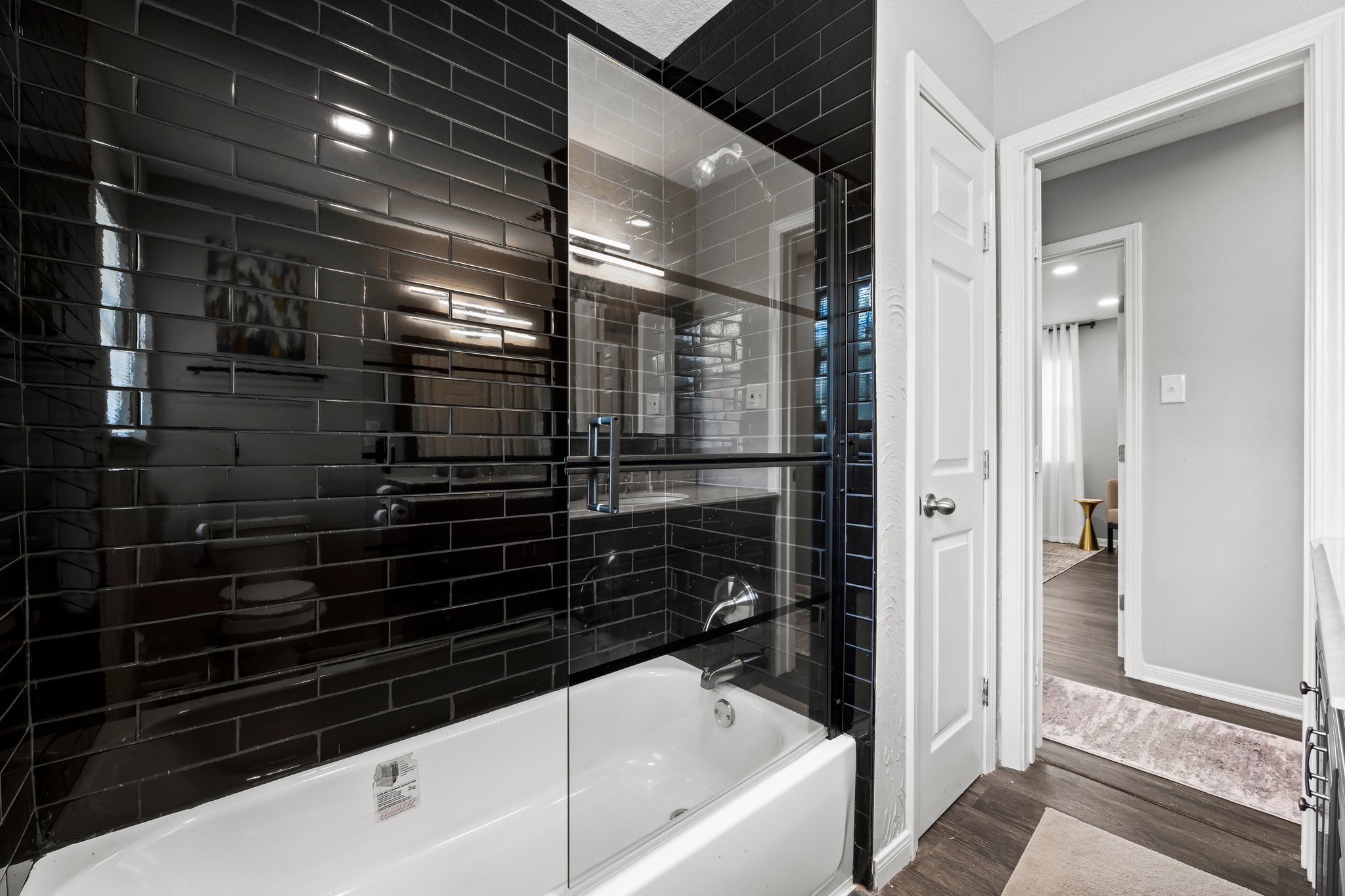 7280 Click Drive Beaumont, TX 77708 - Photo 23 of 39 Modern bathroom featuring sleek black subway tiles, a glass-enclosed bathtub, and an adjacent hallway leading to a stylish room with wood flooring.