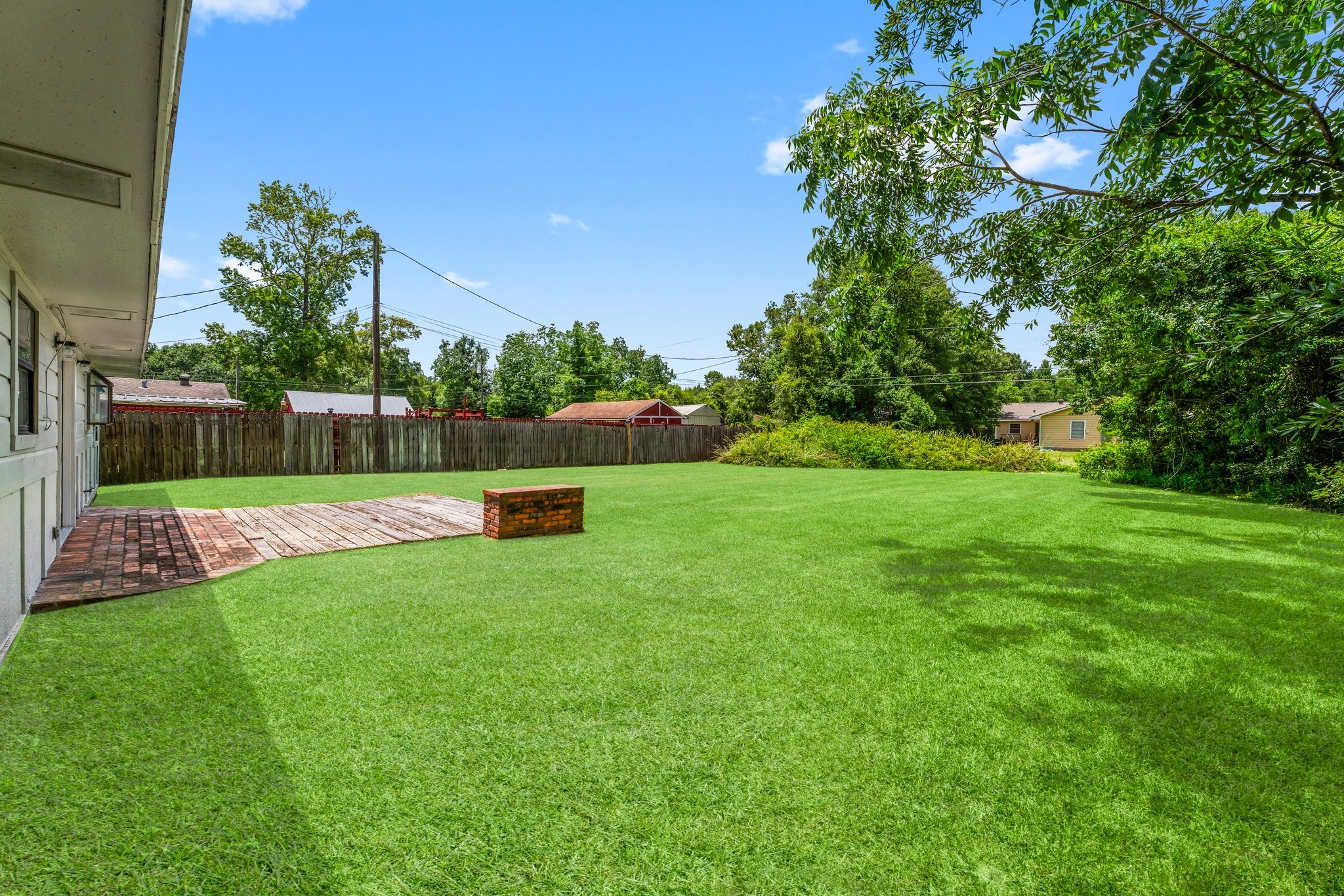 7280 Click Drive Beaumont, TX 77708 - Photo 24 of 39 Spacious backyard with a large grassy area, bordered by a wooden fence. Features a patio with brick pavers and a small raised brick structure, ideal for outdoor activities. Surrounded by trees, offering privacy and a natural feel.