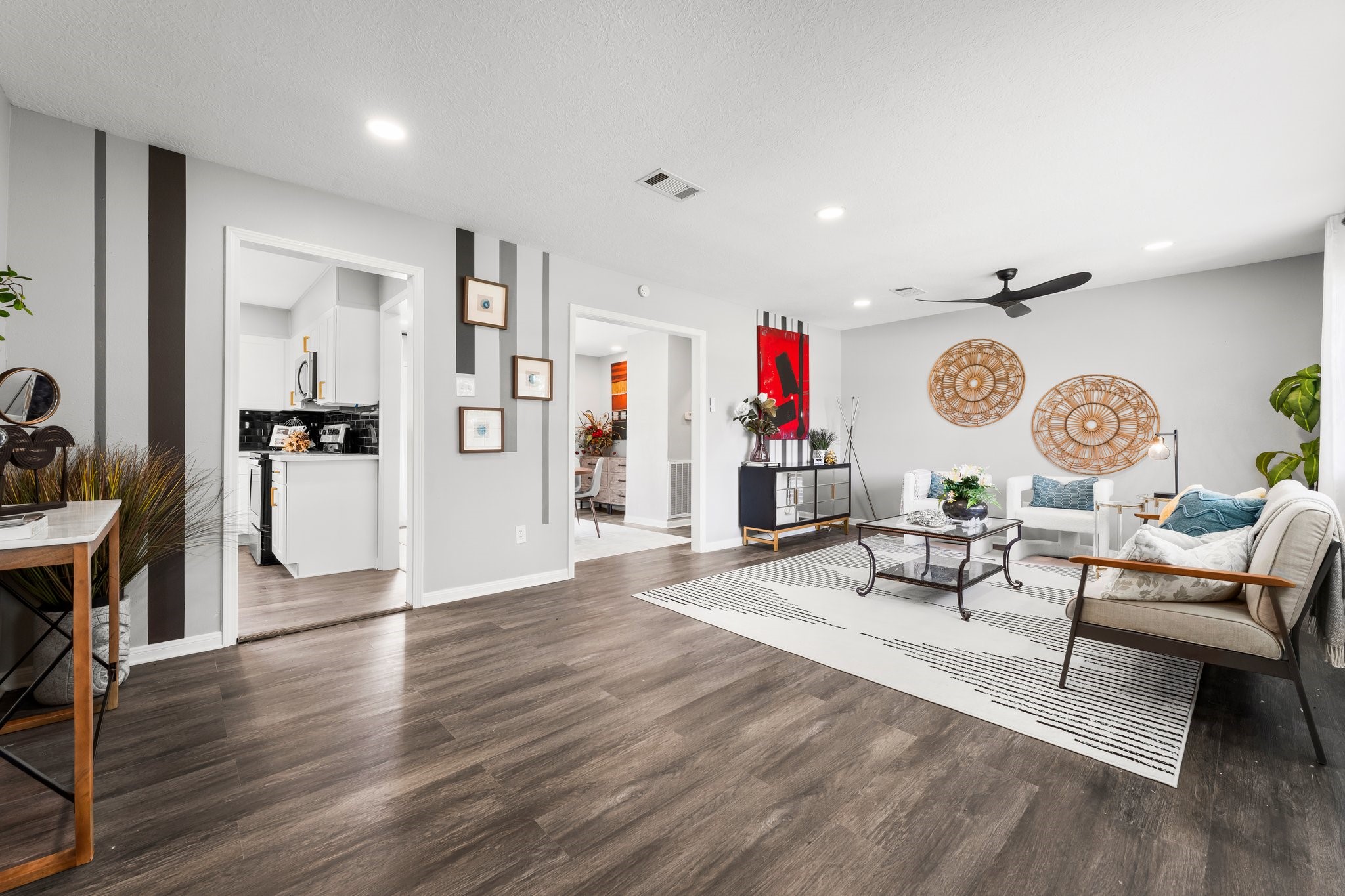 7280 Click Drive Beaumont, TX 77708 - Photo 39 of 39 This photo showcases a modern living room with an open floor plan, featuring wood flooring, stylish decor, and ample natural light. The space is inviting, with a neutral color palette, contemporary furniture, and artistic wall accents. A kitchen and dining area are visible through open doorways, enhancing the spacious feel.