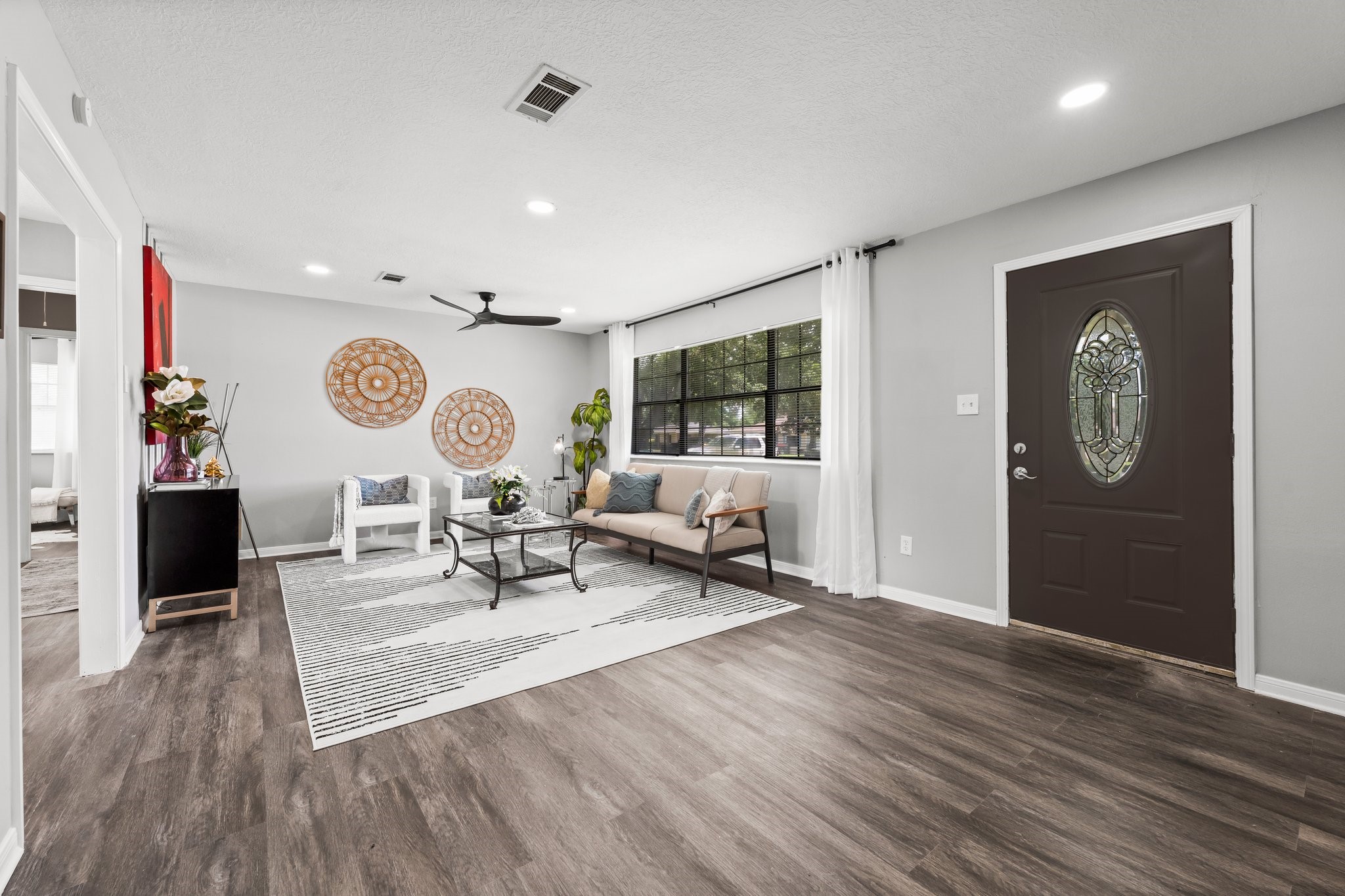 7280 Click Drive Beaumont, TX 77708 - Photo 6 of 39 This inviting living room features a large window for natural light, modern wood flooring, and neutral walls. It includes a stylish ceiling fan, decorative wall art, and an elegant front door with a glass design. The space is tastefully furnished with a cozy sofa, chairs, and a coffee table, perfect for relaxation or entertaining.