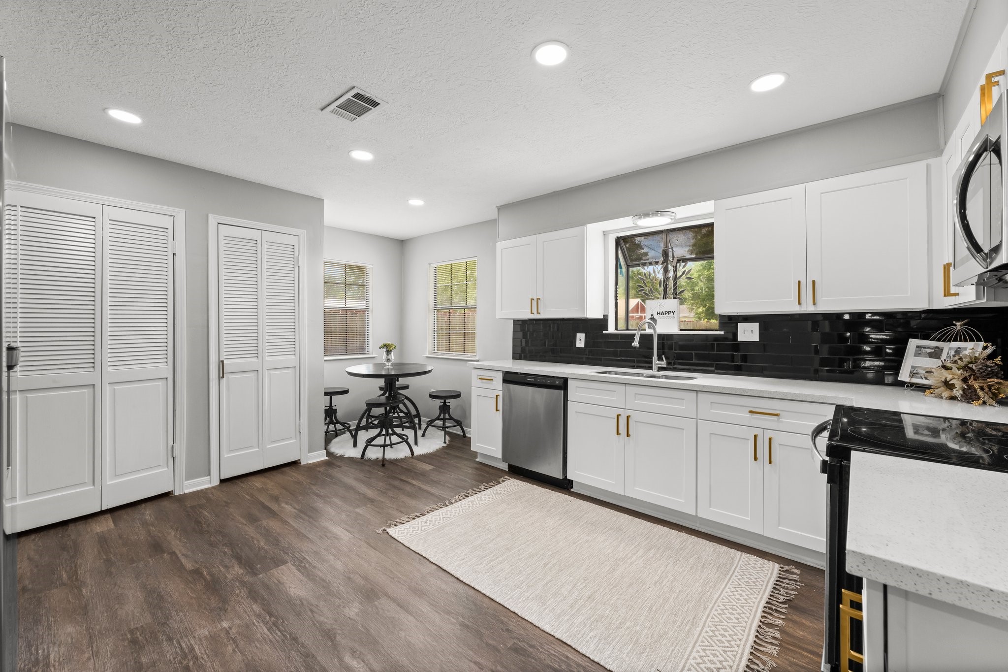 7280 Click Drive Beaumont, TX 77708 - Photo 9 of 39 This kitchen features modern white cabinetry with gold handles, a sleek black backsplash, and stainless steel appliances. The room is brightened by recessed lighting and natural light from the windows, with a cozy breakfast nook and stylish dark wood flooring.