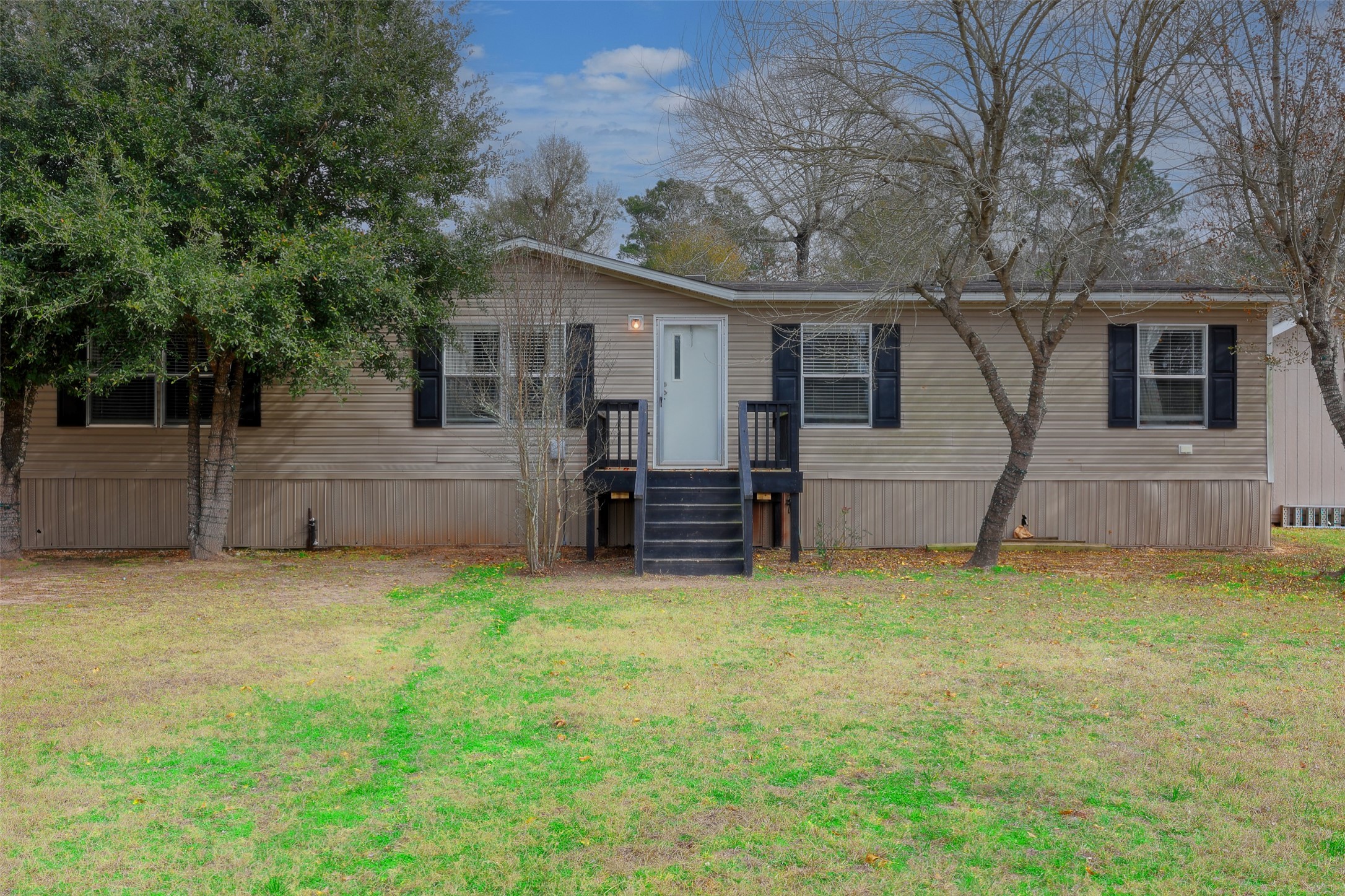 2373 Crockett Martin Road Conroe, TX 77306 - Photo 3 of 34 This photo shows a single-story manufactured home with a neutral exterior and black shutters. It features a small, elevated front porch accessible by stairs and is surrounded by a grassy yard with a few mature trees.