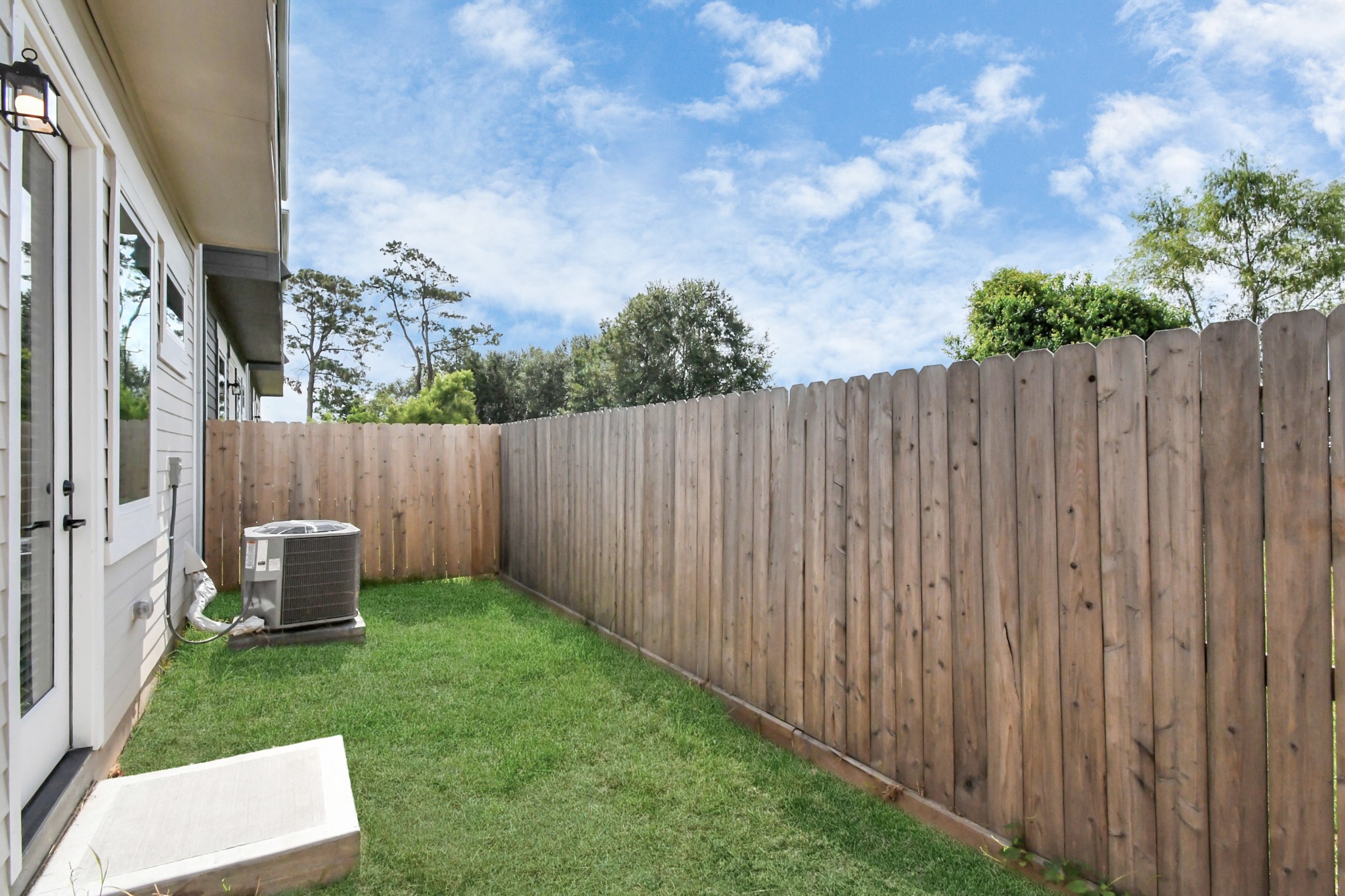 313 Charlock Street Houston, TX 77022 - Photo 29 of 34 This photo shows a small backyard area with a grassy lawn enclosed by a wooden privacy fence. There is an air conditioning unit adjacent to the house, with a clear sky overhead.