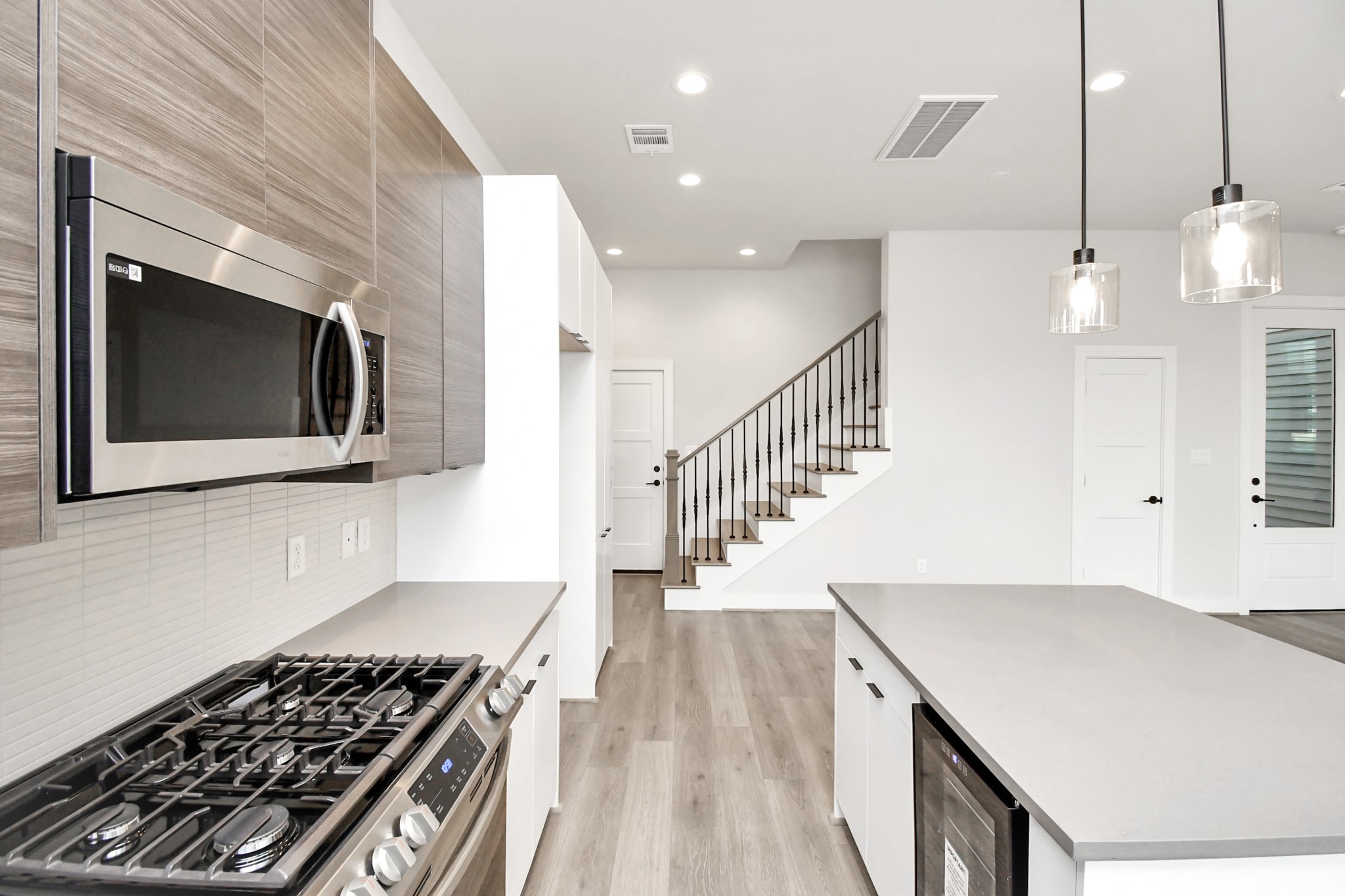 313 Charlock Street Houston, TX 77022 - Photo 10 of 34 Modern kitchen with sleek stainless steel appliances, including a microwave and gas stove. Features a spacious island, pendant lighting, and light wood flooring. Open layout with a view of the staircase and glass door.