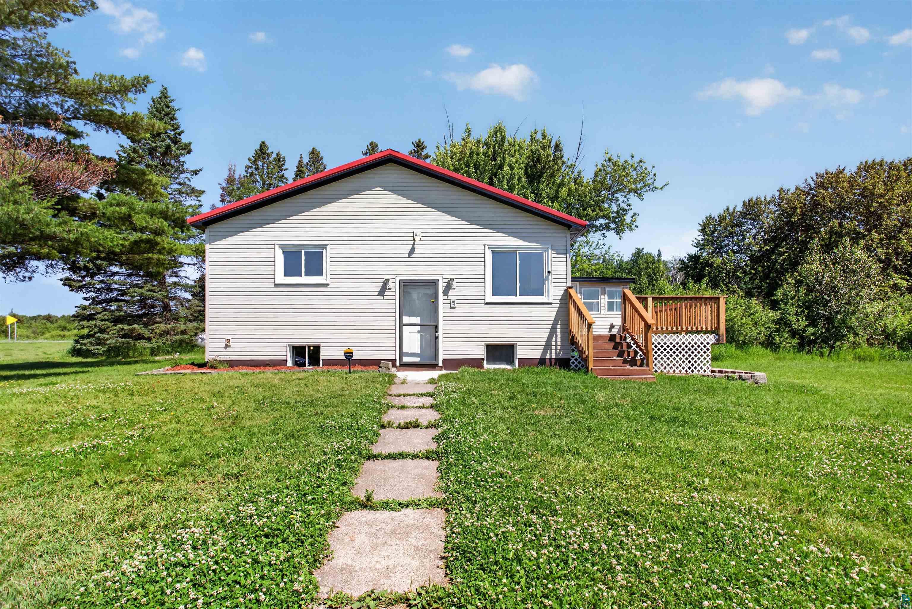 5868 Rice Lake Road Duluth, MN 55803 - Photo 5 of 39 Light-colored horizontal siding exterior with a red roof, featuring a wooden deck with railing, a concrete slab walkway, and multiple window units