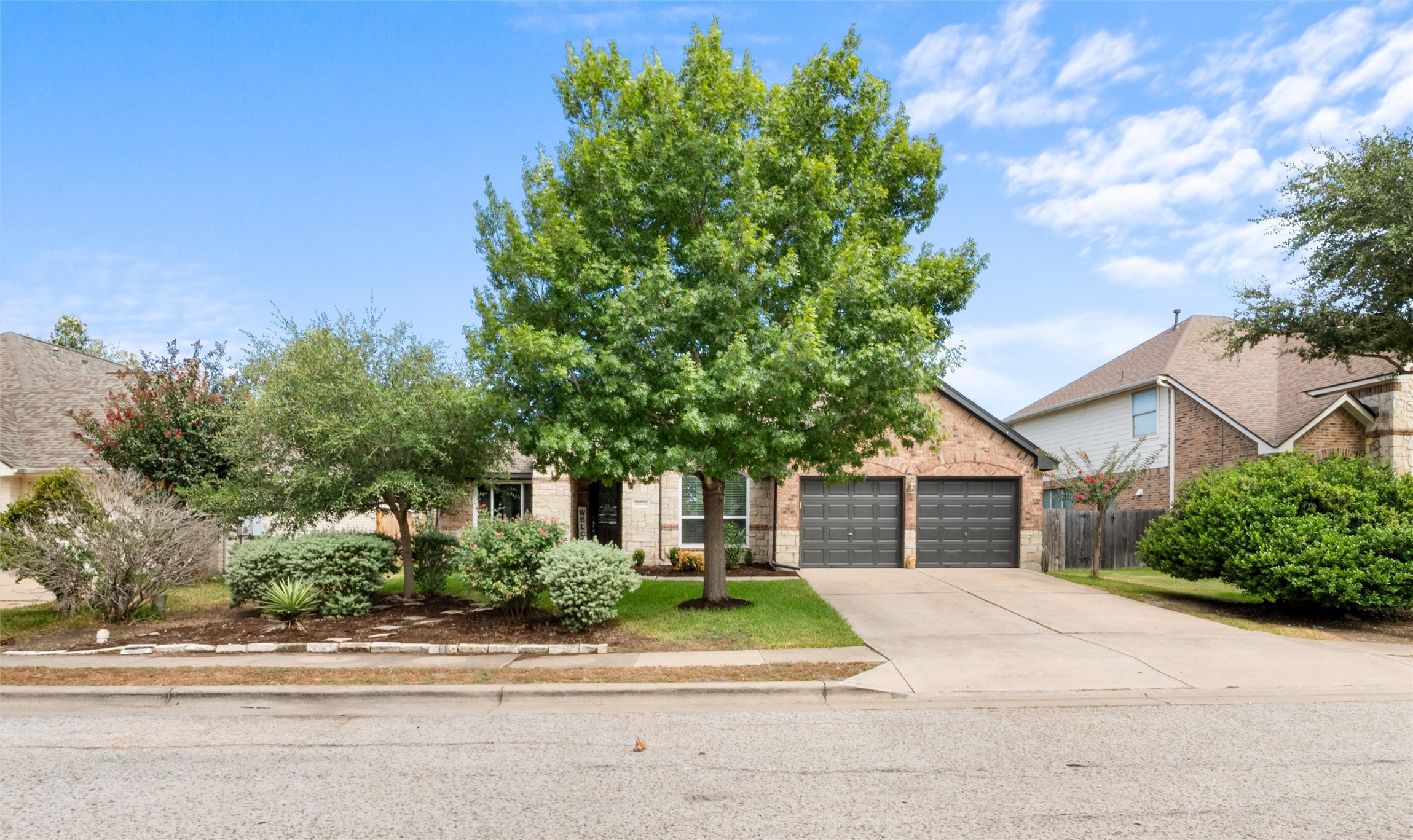 20704 Windmill Ridge Street Pflugerville, TX 78660 - Photo 2 of 34 Mature trees frame a charming brick and stone facade, complemented by a neatly maintained front lawn and driveway.