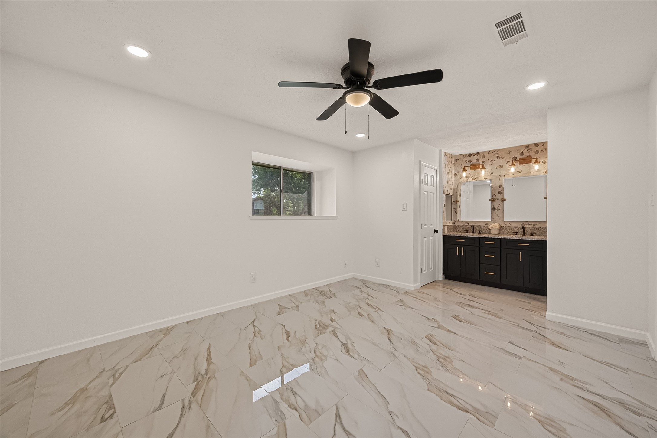 16619 Tibet Road Friendswood, TX 77546 - Photo 13 of 29 This photo showcases a modern bedroom with glossy marble-style tile flooring, a ceiling fan, and recessed lighting. It has a small window for natural light and an open ensuite bathroom featuring a dual-sink vanity with dark cabinetry and decorative lighting.