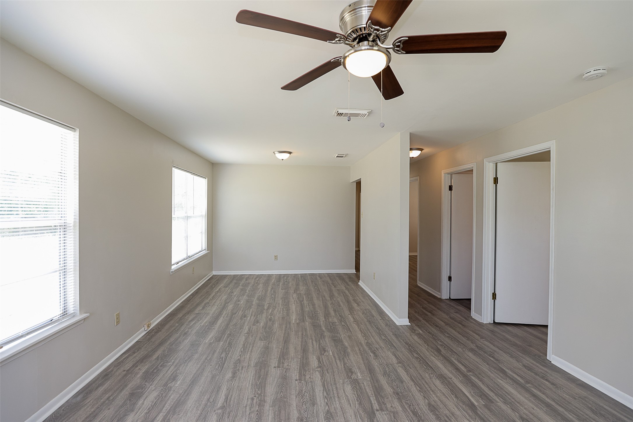 7810 Queen Street Houston, TX 77028 - Photo 3 of 21 This is a spacious, well-lit room featuring modern gray wood-like flooring, a ceiling fan with lighting, neutral wall colors, and multiple windows for natural light. The room leads to a hallway with additional rooms, suggesting a functional layout.