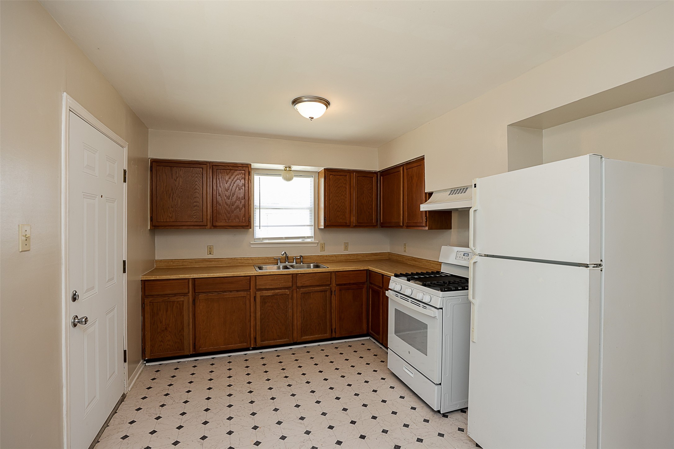 7810 Queen Street Houston, TX 77028 - Photo 7 of 21 This is a functional kitchen with traditional wooden cabinets, white appliances including a refrigerator and a range, a double sink beneath a window, and a patterned vinyl floor.