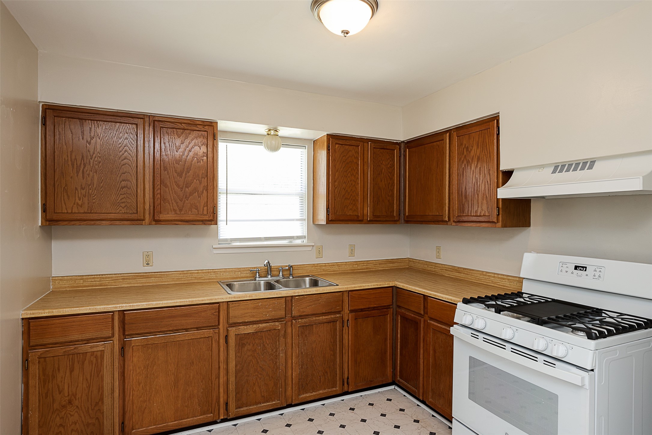 7810 Queen Street Houston, TX 77028 - Photo 8 of 21 This is a compact kitchen featuring traditional wood cabinetry, laminate countertops, a double sink, gas stove, and a built-in overhead exhaust hood. The kitchen has a window for natural light, vinyl tile flooring, and a flush mount ceiling light.