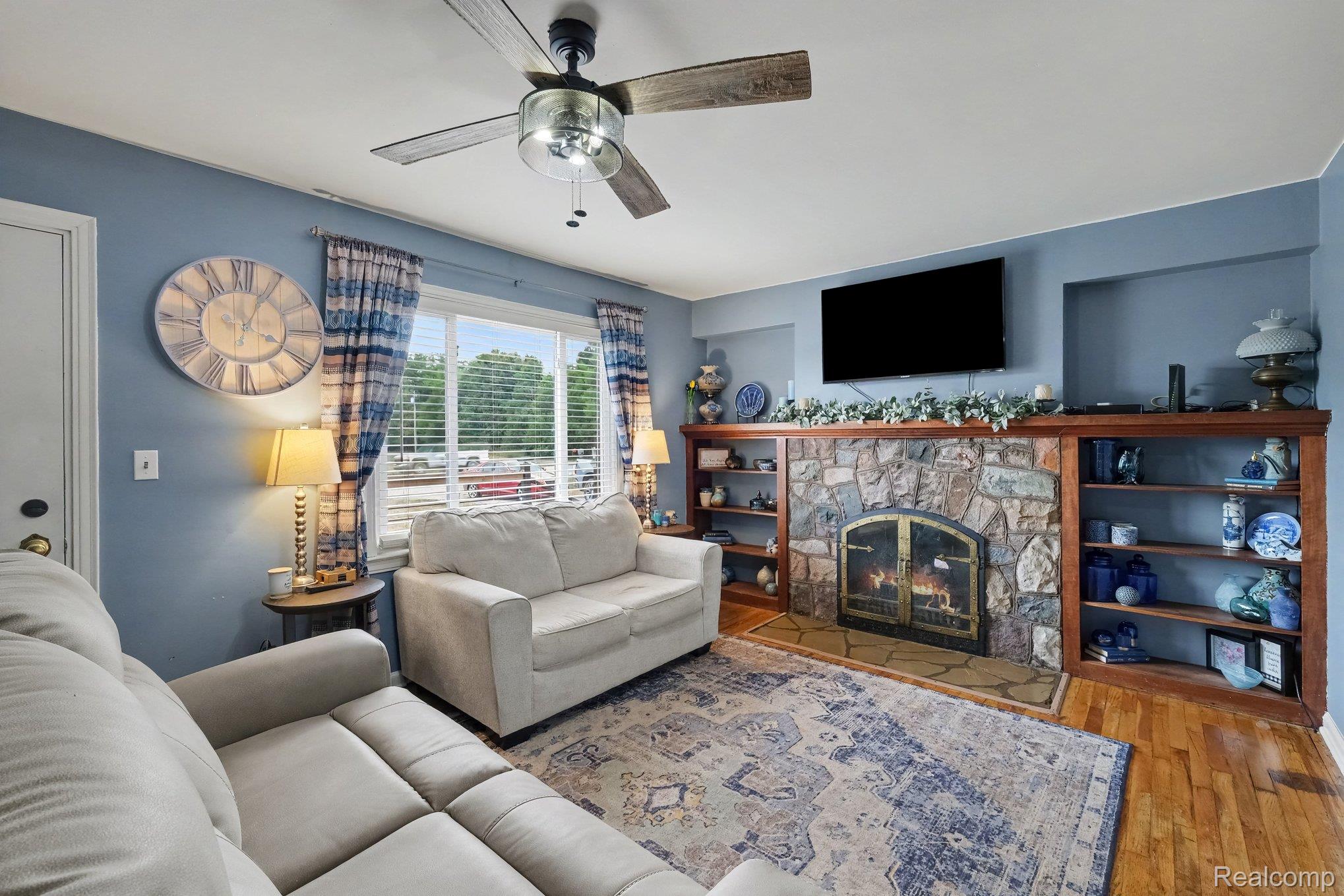 5409 Baldwin Road Swartz Creek, MI 48473 - Photo 2 of 27 Stone-clad fireplace featuring a hearth and built-in shelving, wood-finish flooring, and a ceiling fan with integrated lighting