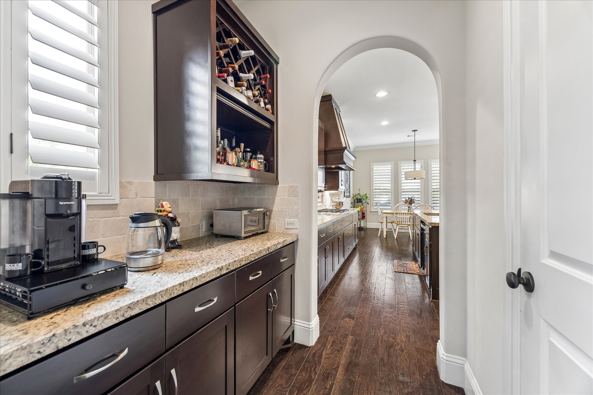 3 Tiburon Court Spring, TX 77389 - Photo 10 of 37 Butler's pantry with dark wood cabinets, granite countertops, and a built-in wine rack. The space features hardwood floors and an arched doorway leading to a bright dining area with large windows and elegant light fixtures.