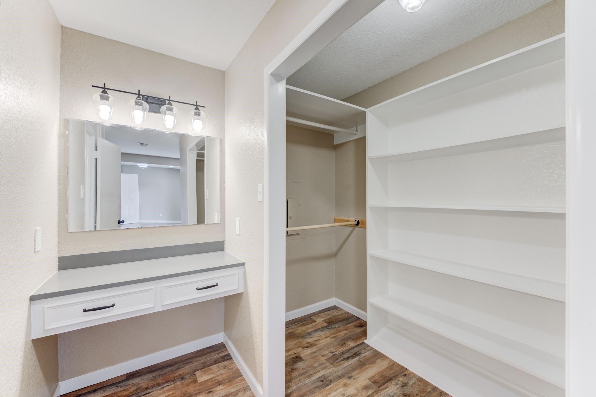 305 Yaupon Street Sweeny, TX 77480 - Photo 19 of 22 This photo showcases a bright walk-in closet with ample shelving and hanging space, adjacent to a vanity area with a large mirror and modern lighting. The flooring is a warm, wood-like finish, providing a cozy yet functional dressing area.