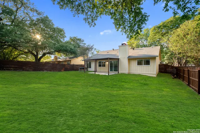 a view of a white house in front of a big yard with large trees and wooden fence